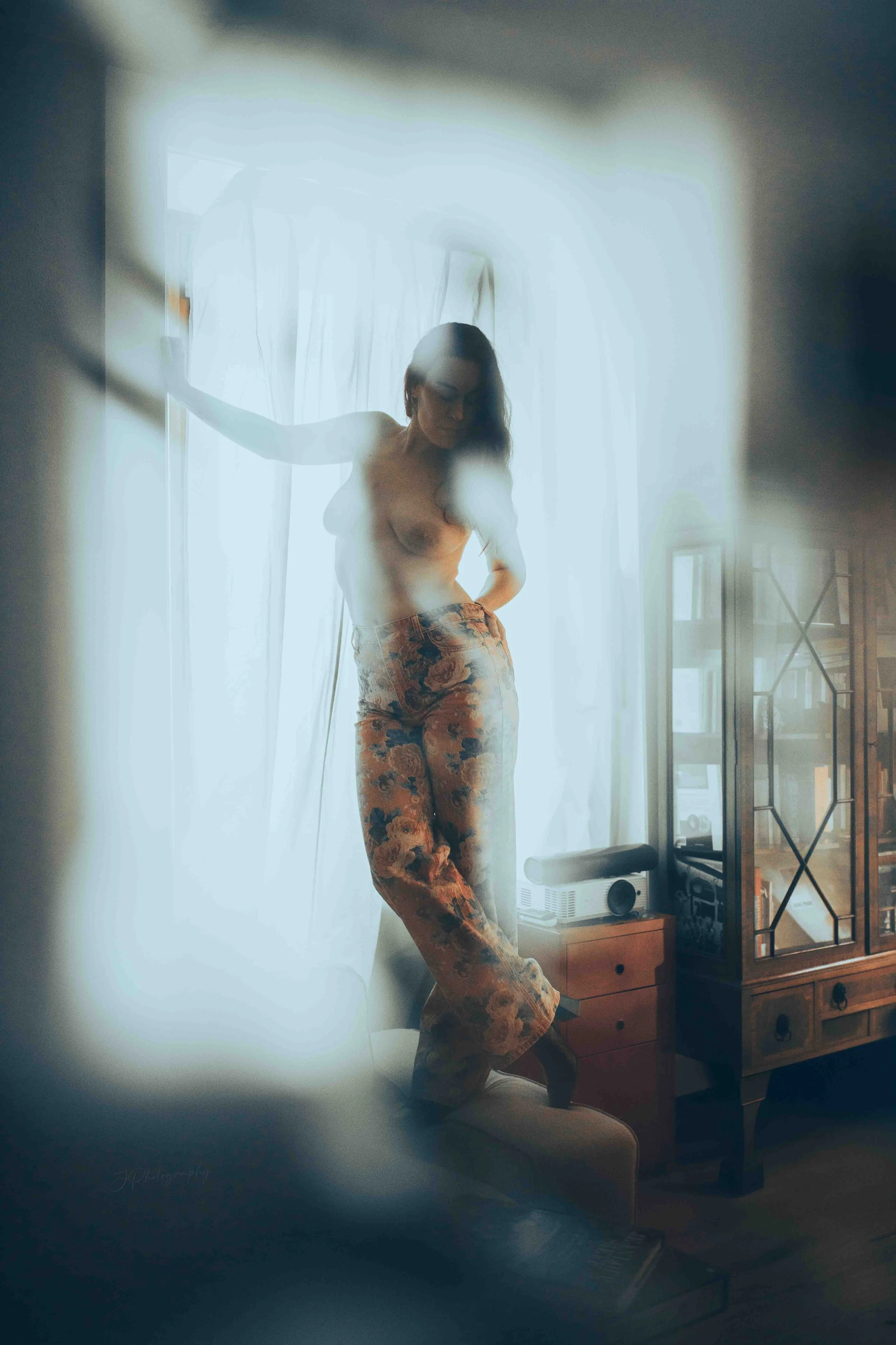Femme nue portant un pantalon à motifs, posant dans une pièce lumineuse avec des rideaux blancs, vue à travers une surface floue ou réfléchissante.