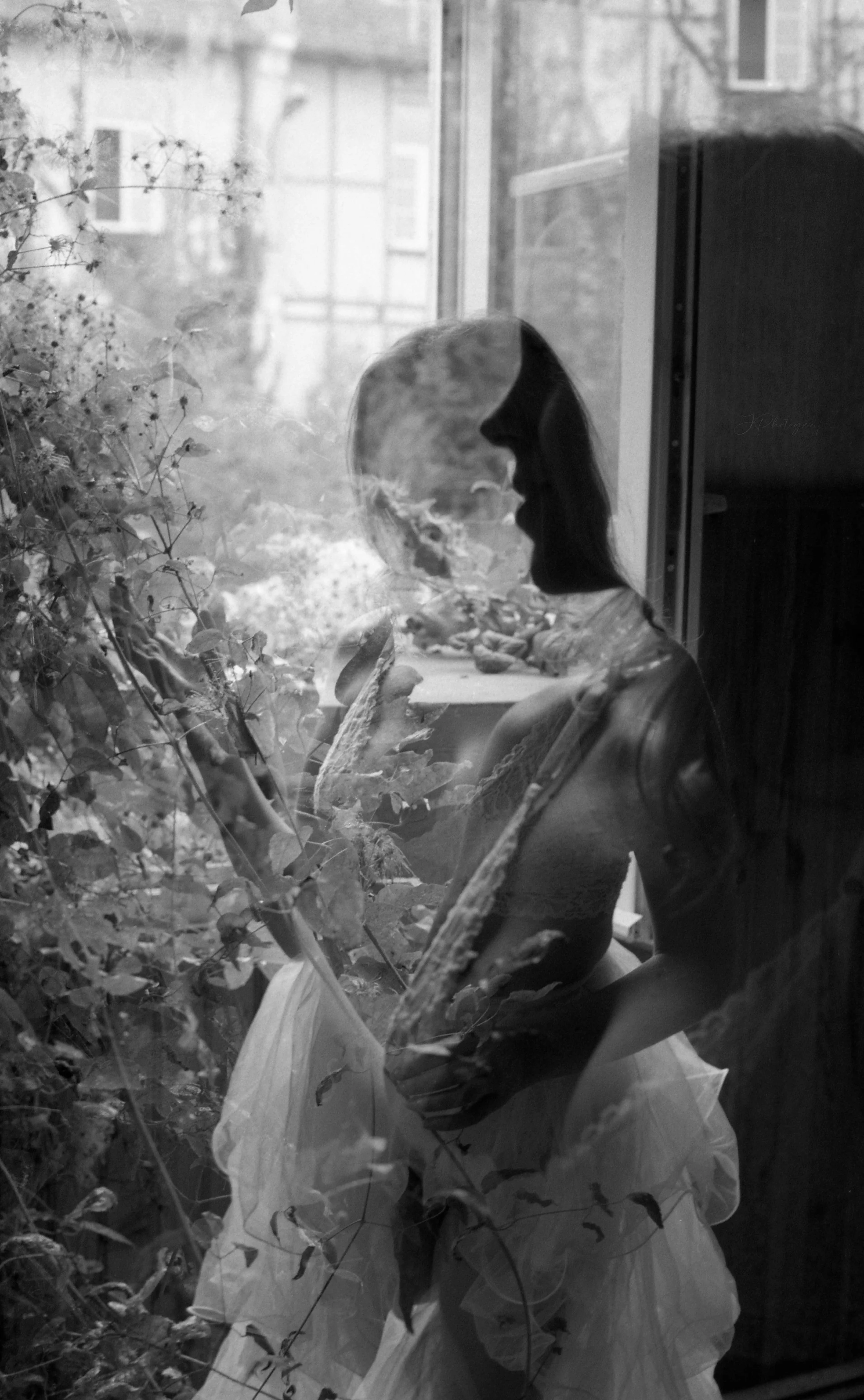 Une femme en lingerie et jupe, vue à travers une fenêtre, avec des plantes et un intérieur en arrière-plan.