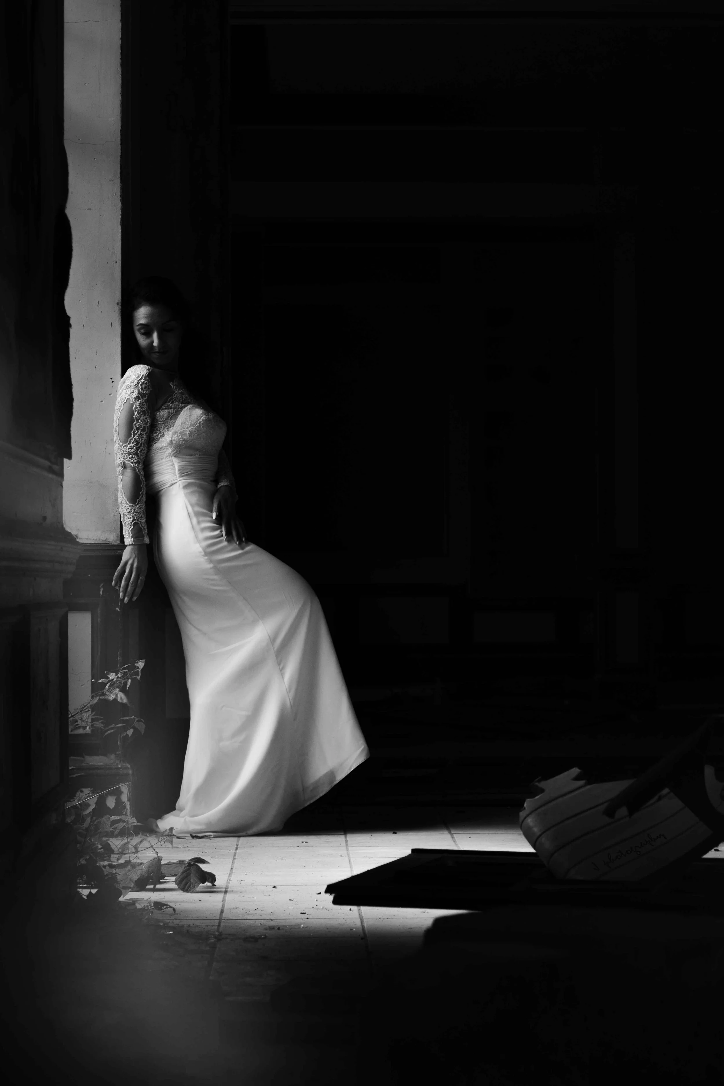 Une femme en robe de mariée se repose contre un mur dans une pièce sombre, avec des feuilles mortes au sol et un piano à gauche.