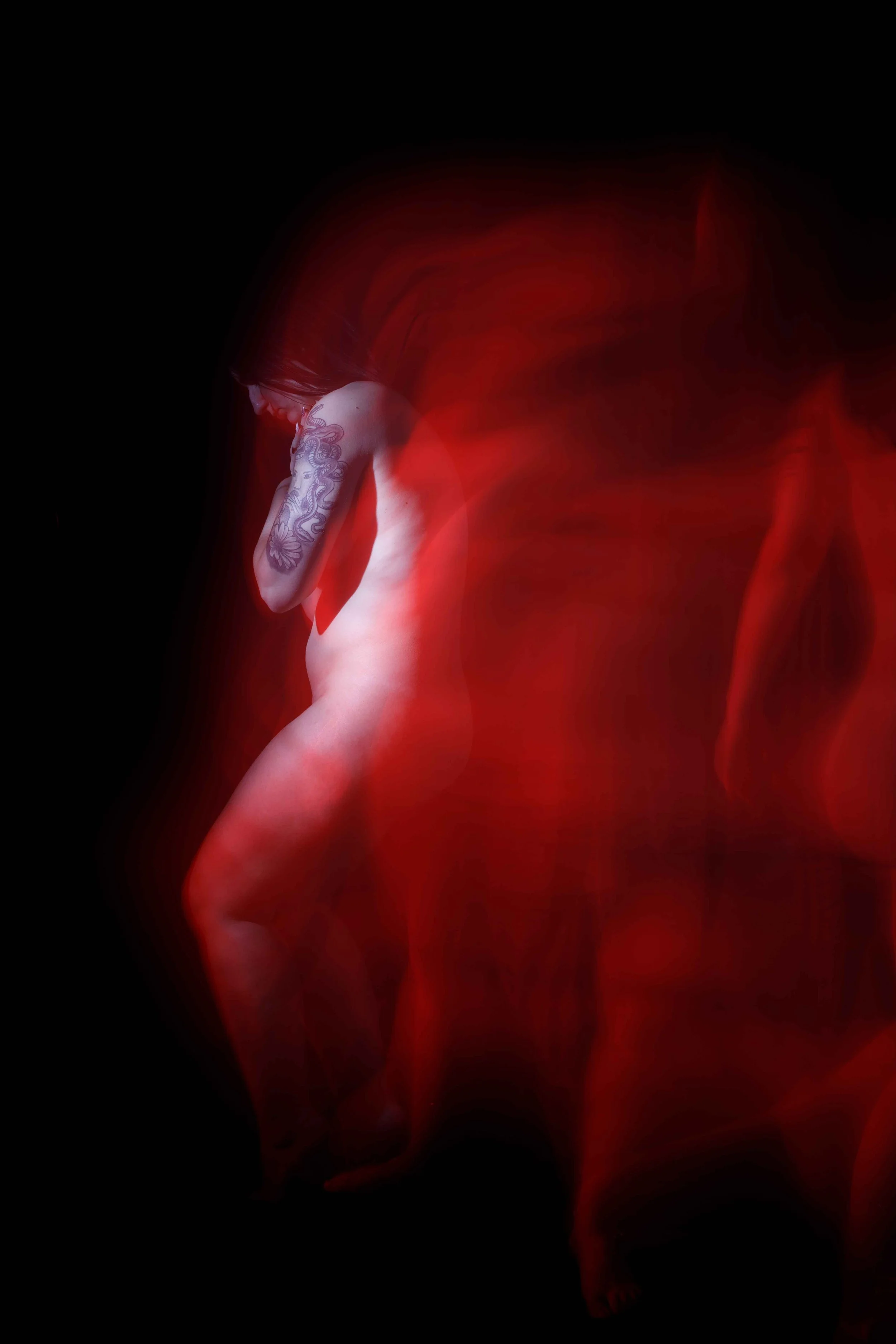 Une femme nue avec un tatouage sur le bras gauche, posant sur un fond noir avec un effet de lumière rouge flottante autour d'elle.
