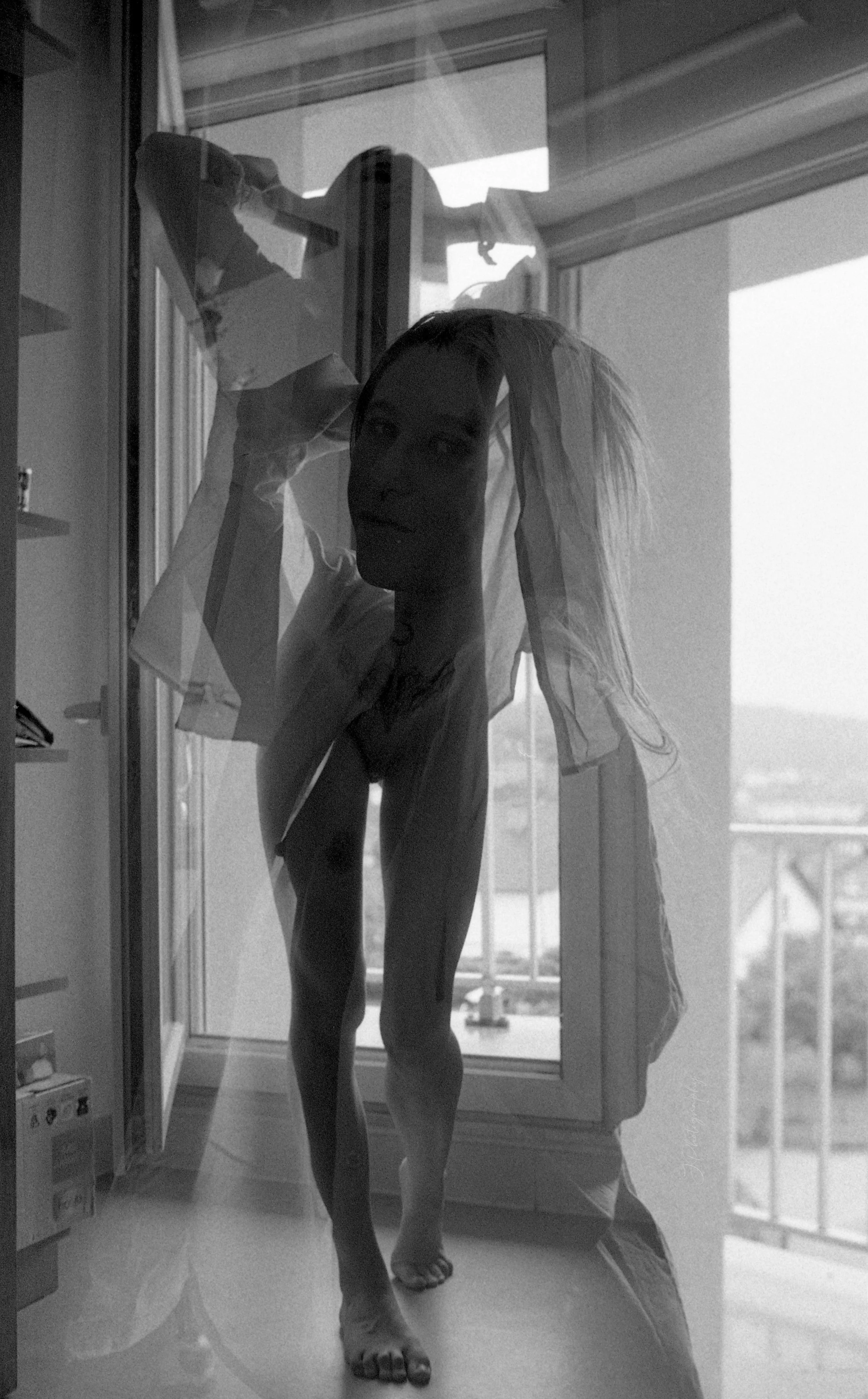 Une jeune femme se tient devant une fenêtre, regardant à l'extérieur, avec un voile transparent couvrant son visage. La scène est en noir et blanc.