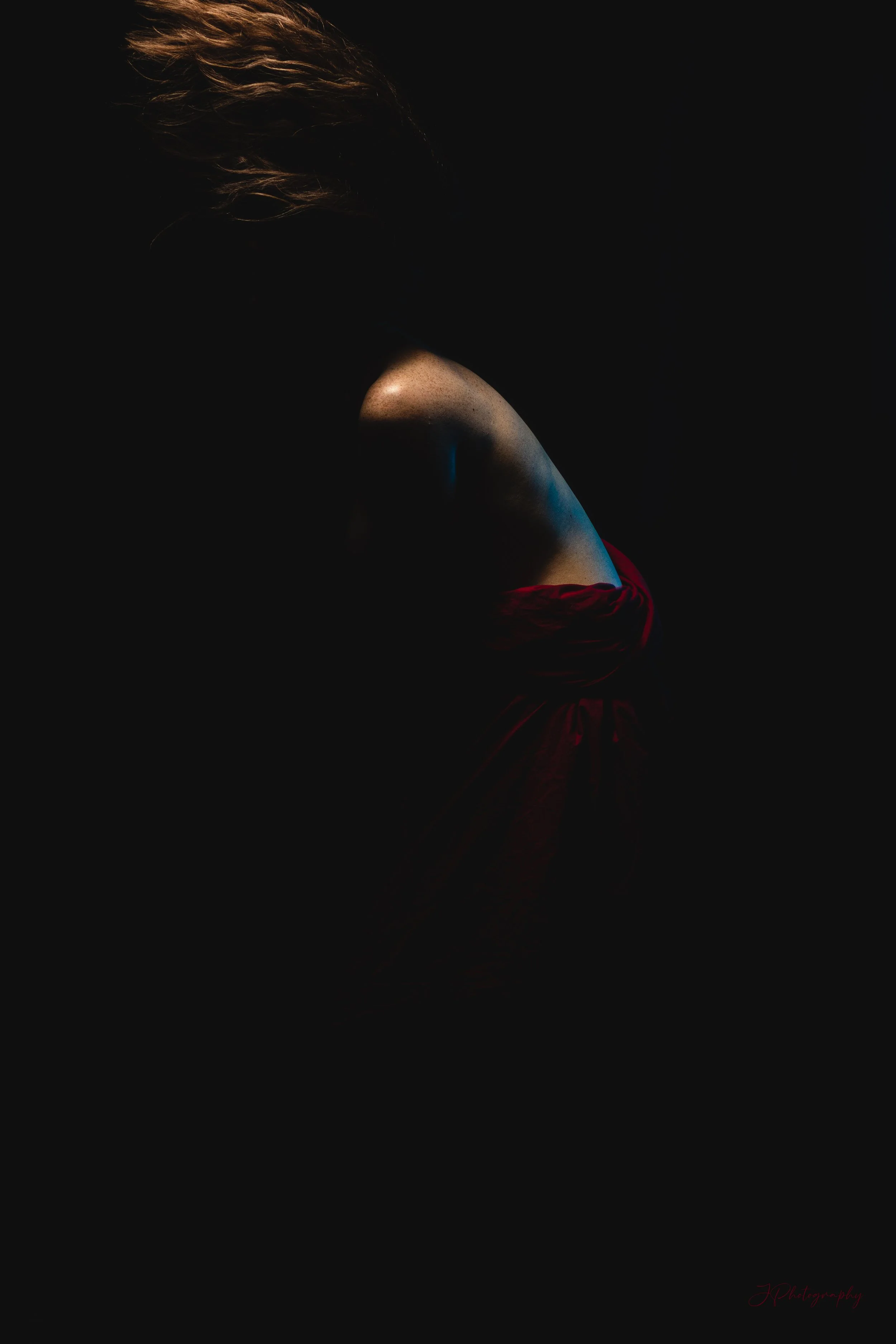 Portrait sombre d'une femme partiellement cachée dans l'ombre, avec un seul épaule visible et un foulard rouge autour du cou.