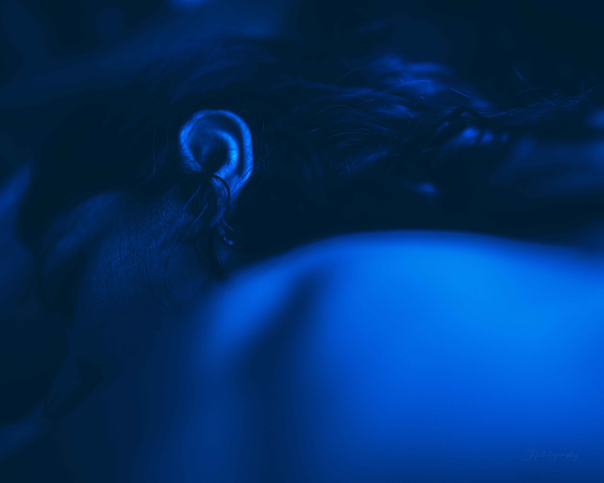 Obscure close-up of a human ear illuminated by blue light, with hair visible around it, creating a mysterious atmosphere.