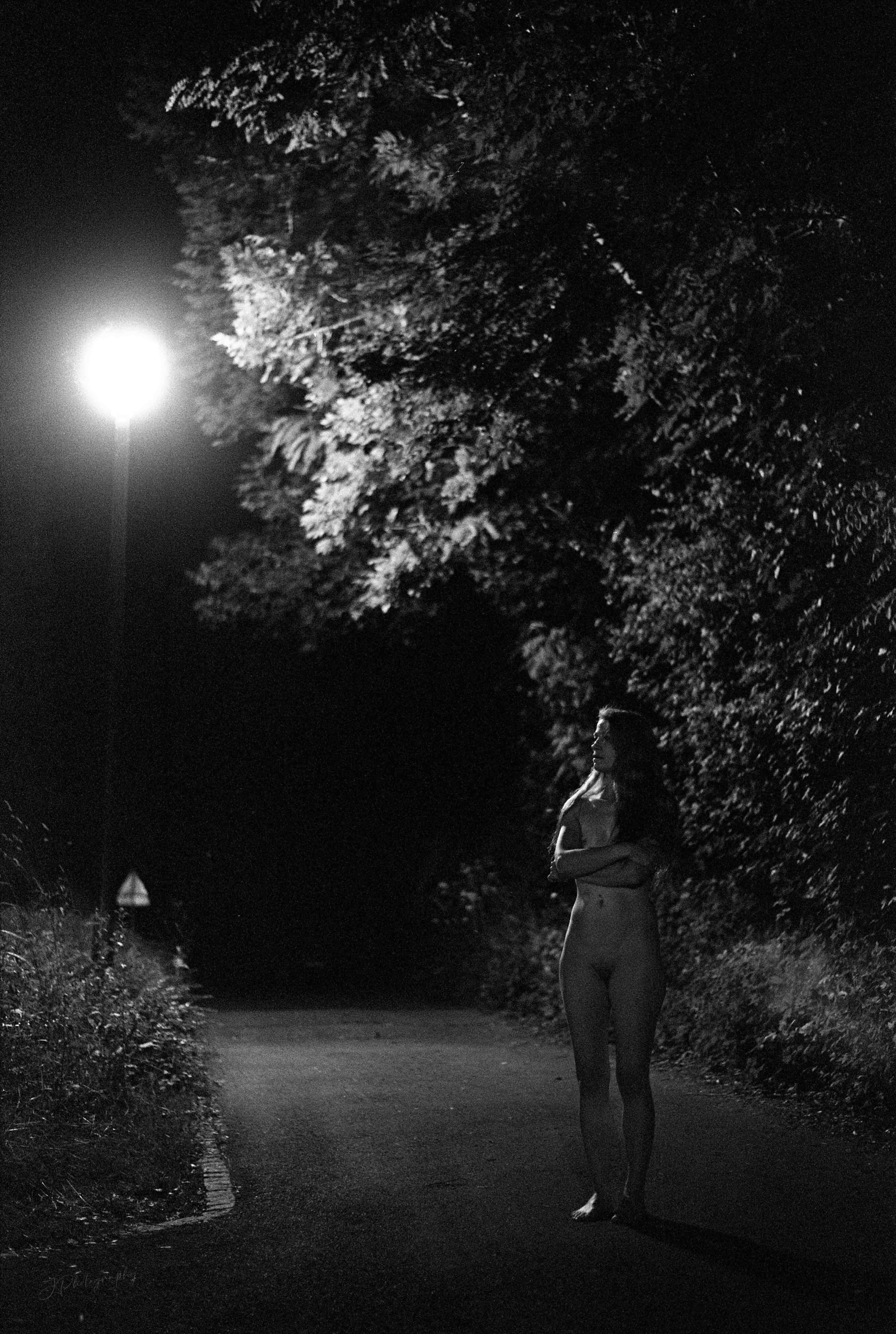 Une femme nue se tient sur un chemin sombre sous un ciel nocturne, éclairée par la lumière d'une grande lune. Elle a des cheveux longs et regarde vers le ciel.