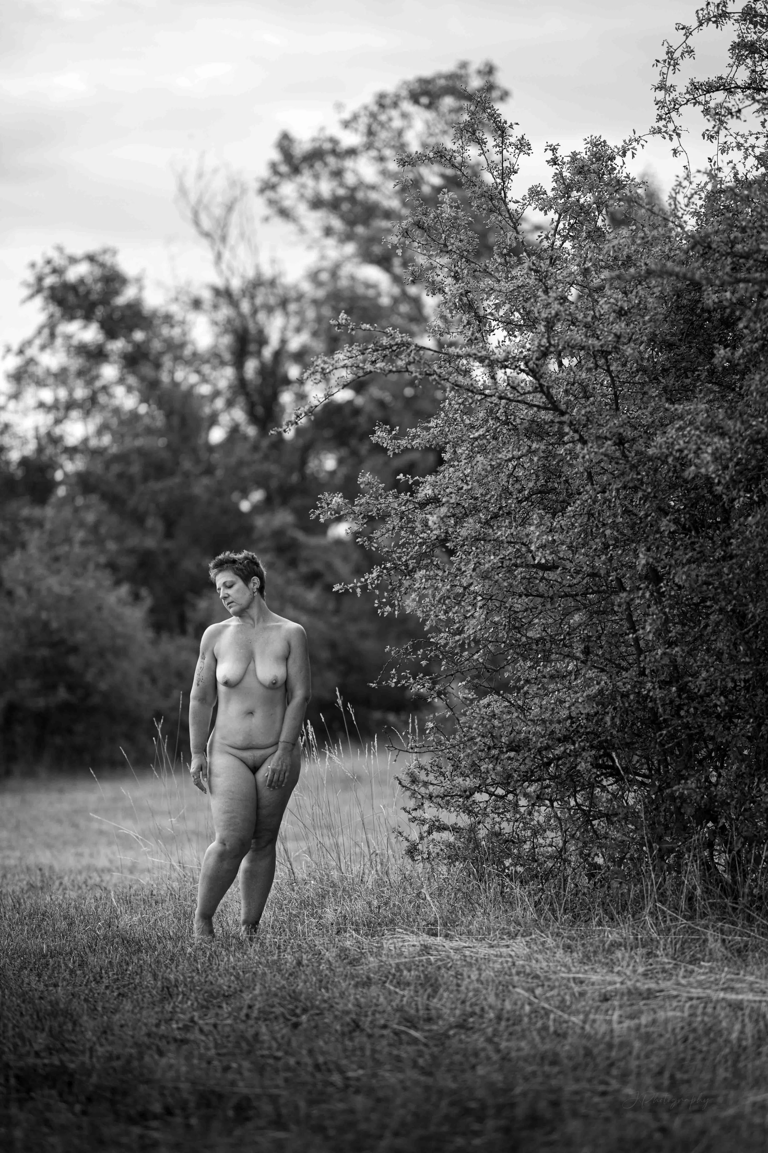 Une femme nue marche seule dans un champ herbeux avec des arbres en arrière-plan, en noir et blanc.