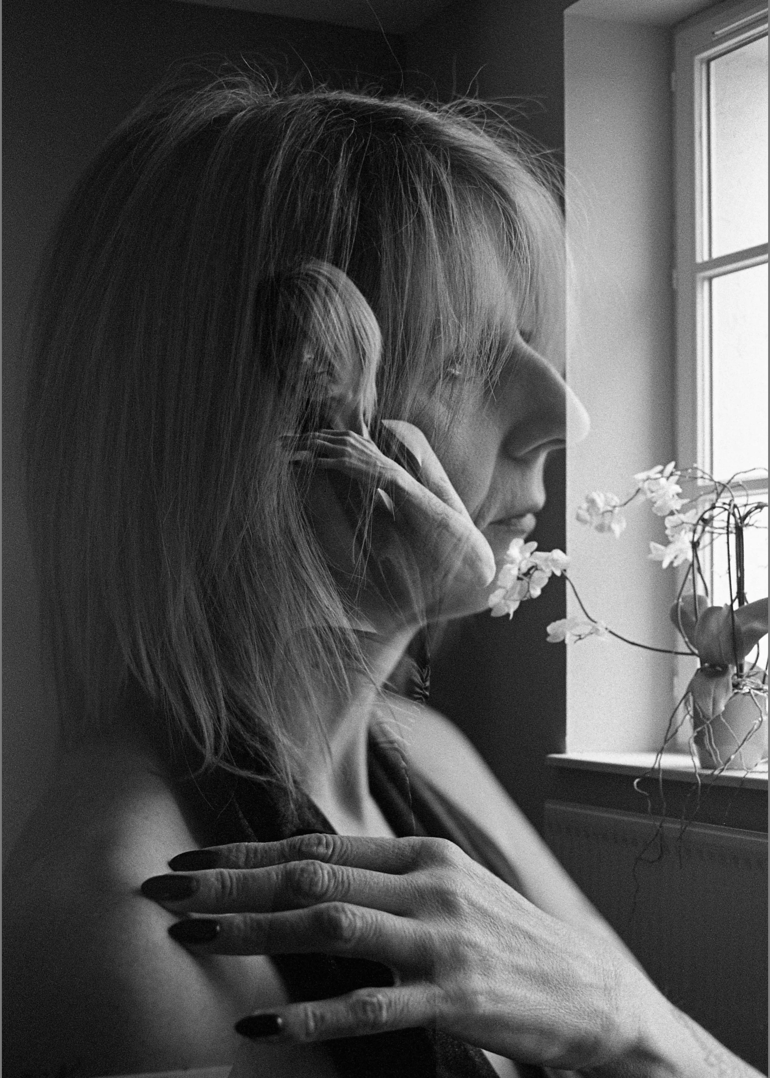Une femme avec des cheveux longs regarde par une fenêtre, sa main posée sur son épaule, en noir et blanc.