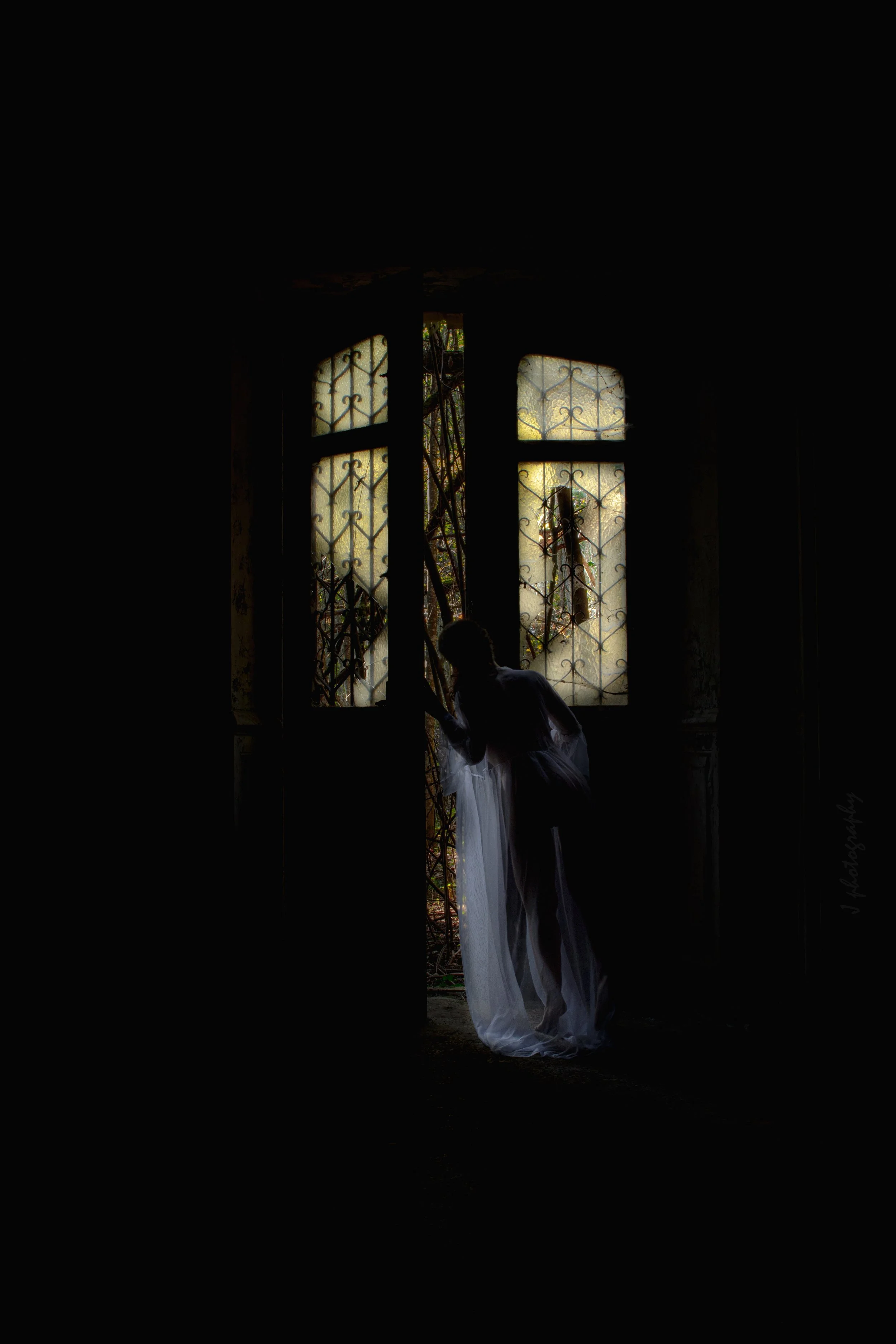 Une silhouette de femme en robe blanche regarde par une porte cassée avec des fenêtres à verre dépoli et des grilles en métal à l'intérieur d'une pièce sombre.
