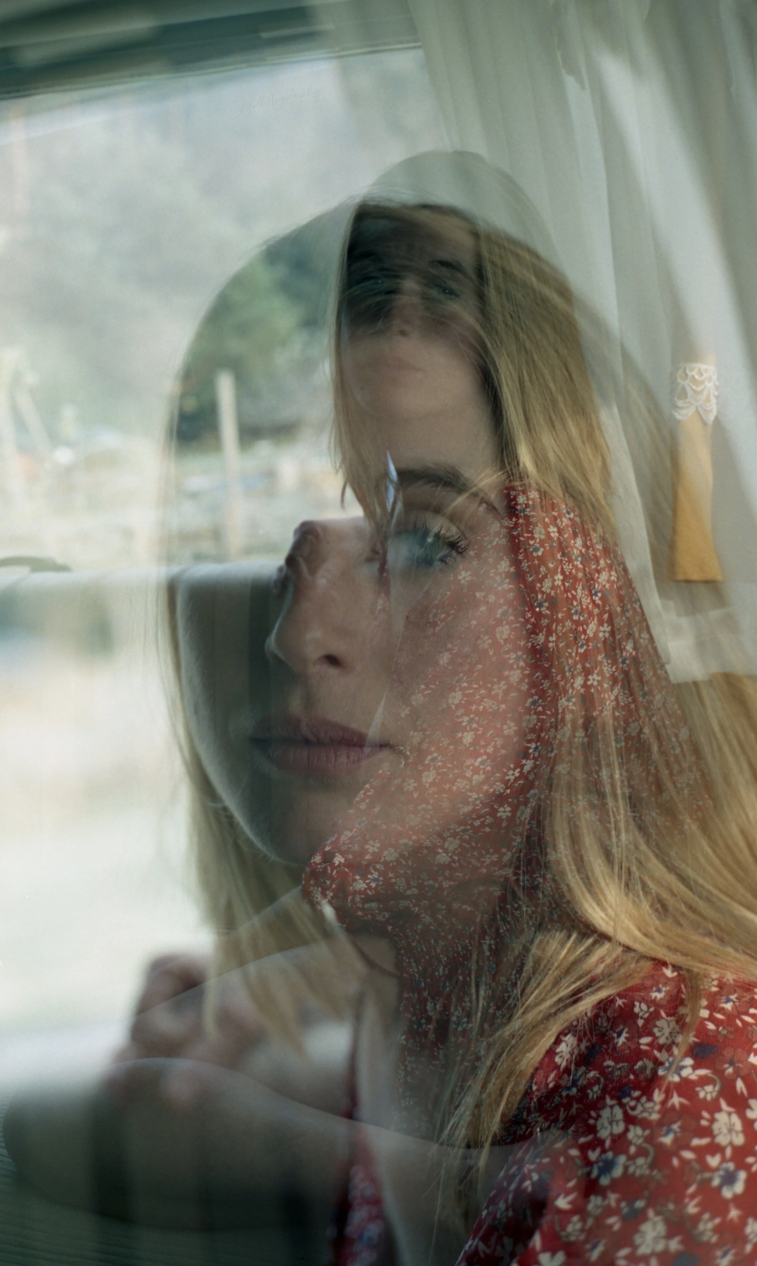 Une femme blonde avec des cheveux longs, porte une robe à motif floral rouge, vue à travers une vitre, avec un reflet superposé d'une autre image de son visage en gros plan.