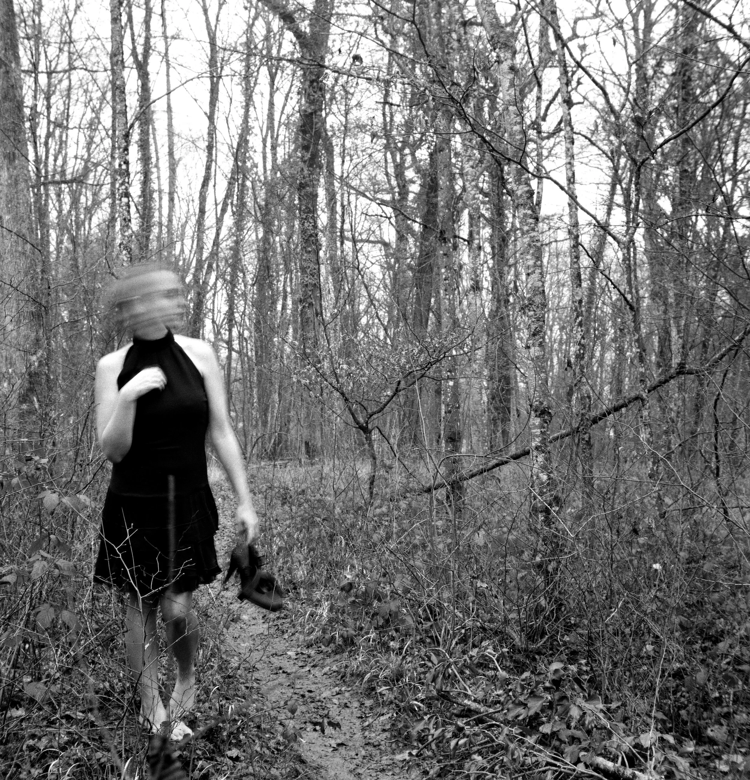 Une femme vêtue d'une robe noire marche dans une forêt, la photo est floue et en noir et blanc.