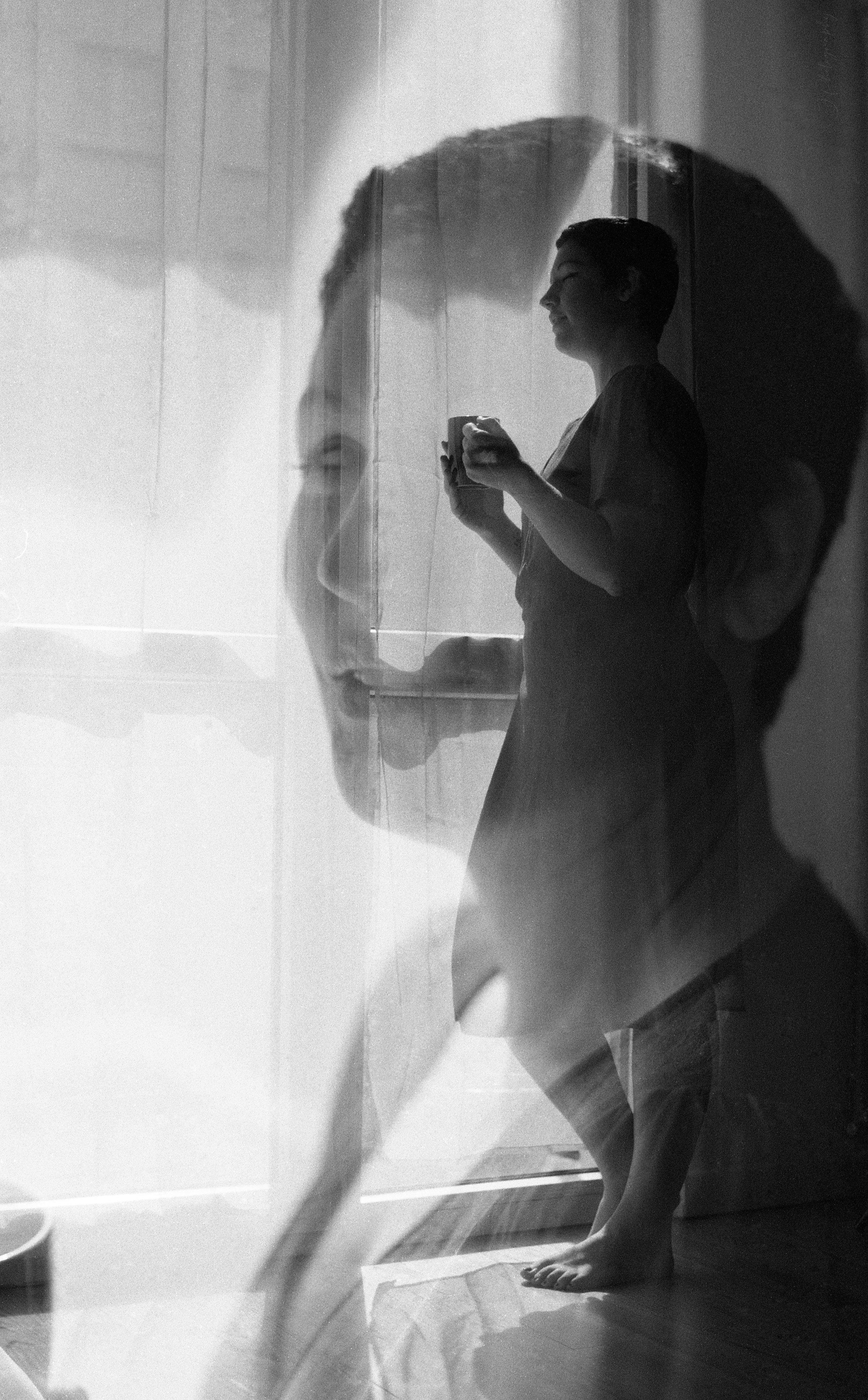 Double exposition argentique. Une femme souriante debout dans une salle de bain, tenant une tasse, avec une double exposition d'une autre personne dans une fenêtre ou un miroir