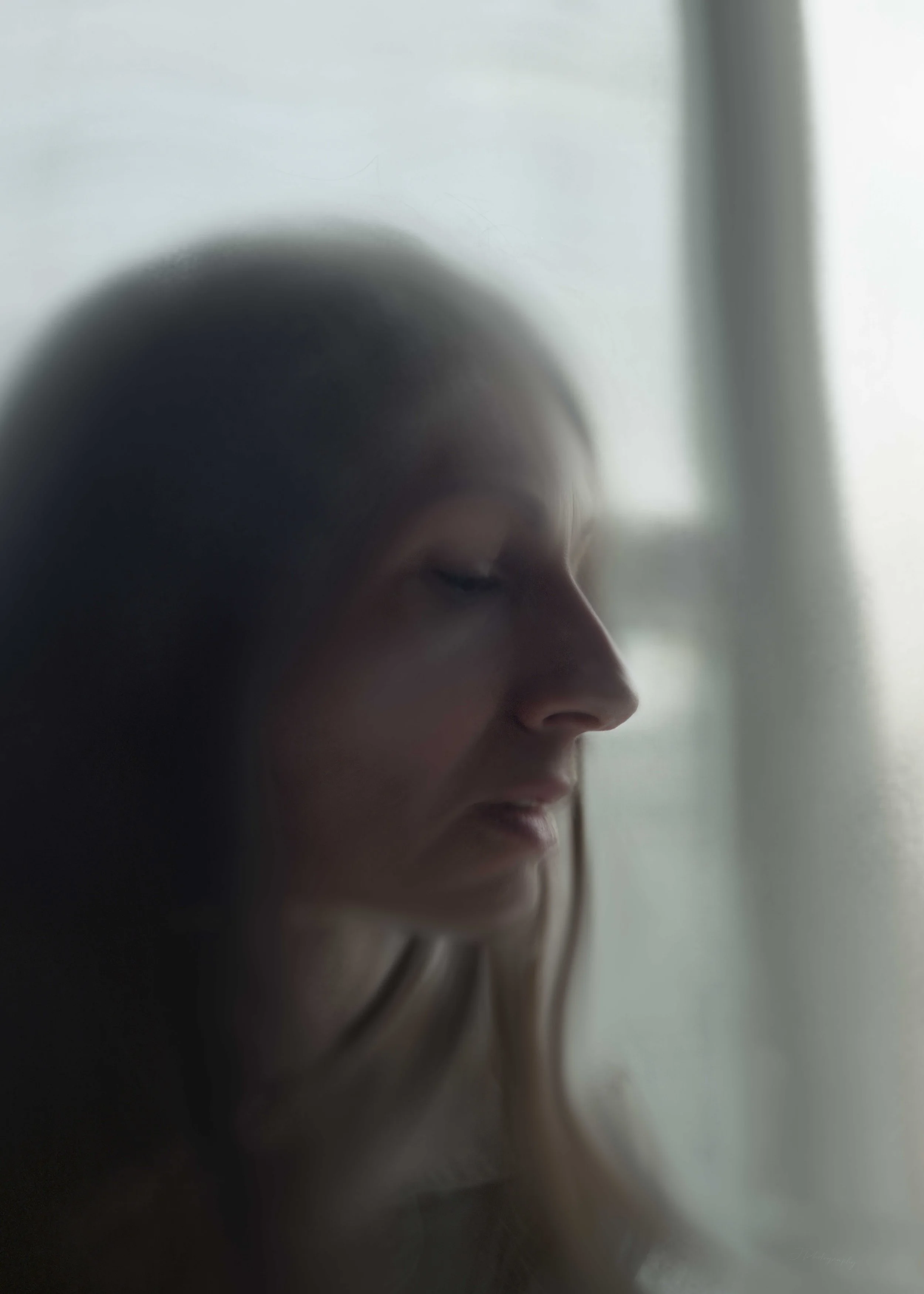 Une femme aux cheveux longs regarde doucement par une fenêtre, avec une lumière naturelle tamisée.