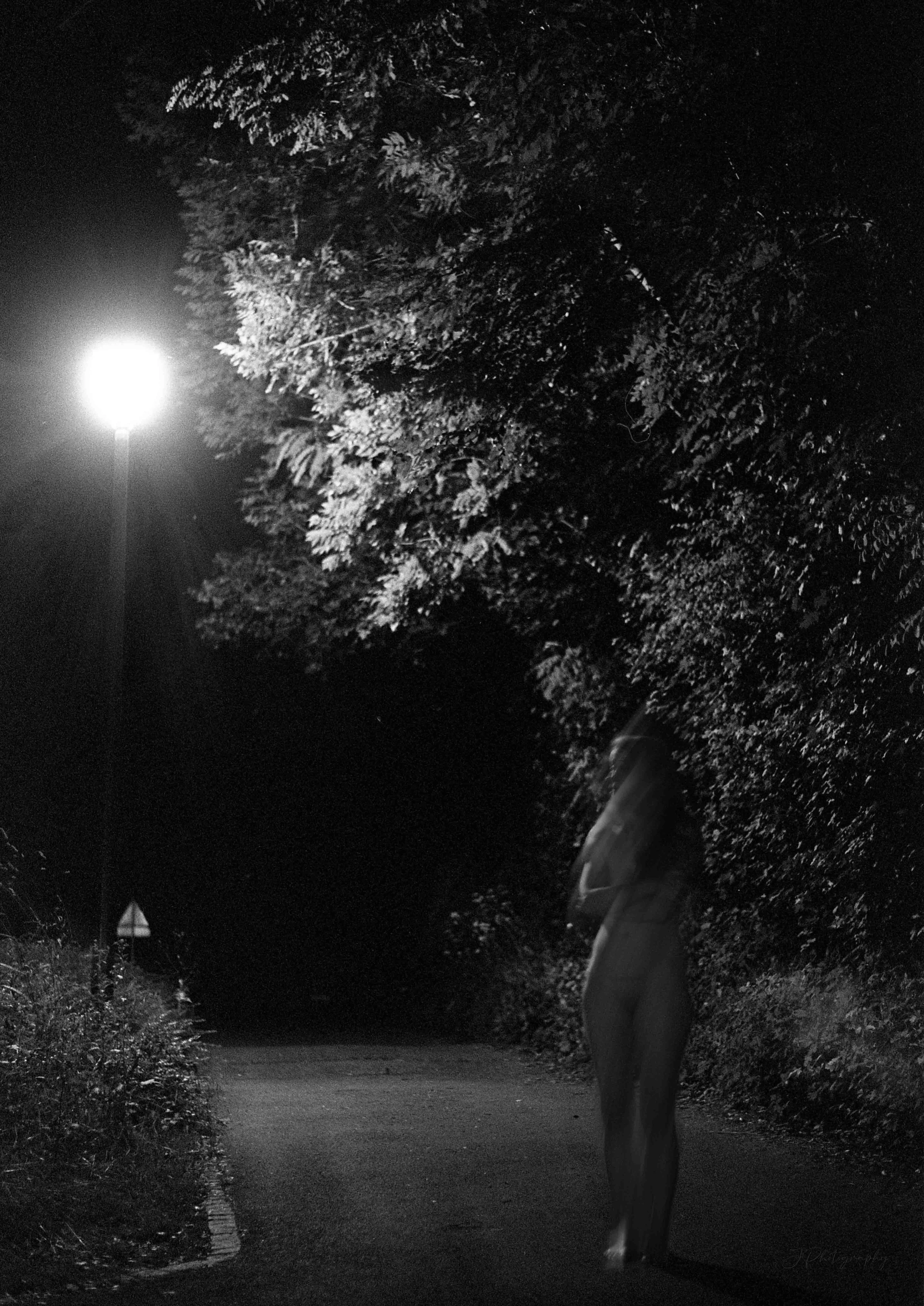 Photo en noir et blanc d'une personne qui marche sur un chemin bordé d’arbres, éclairé par un lampadaire la nuit.