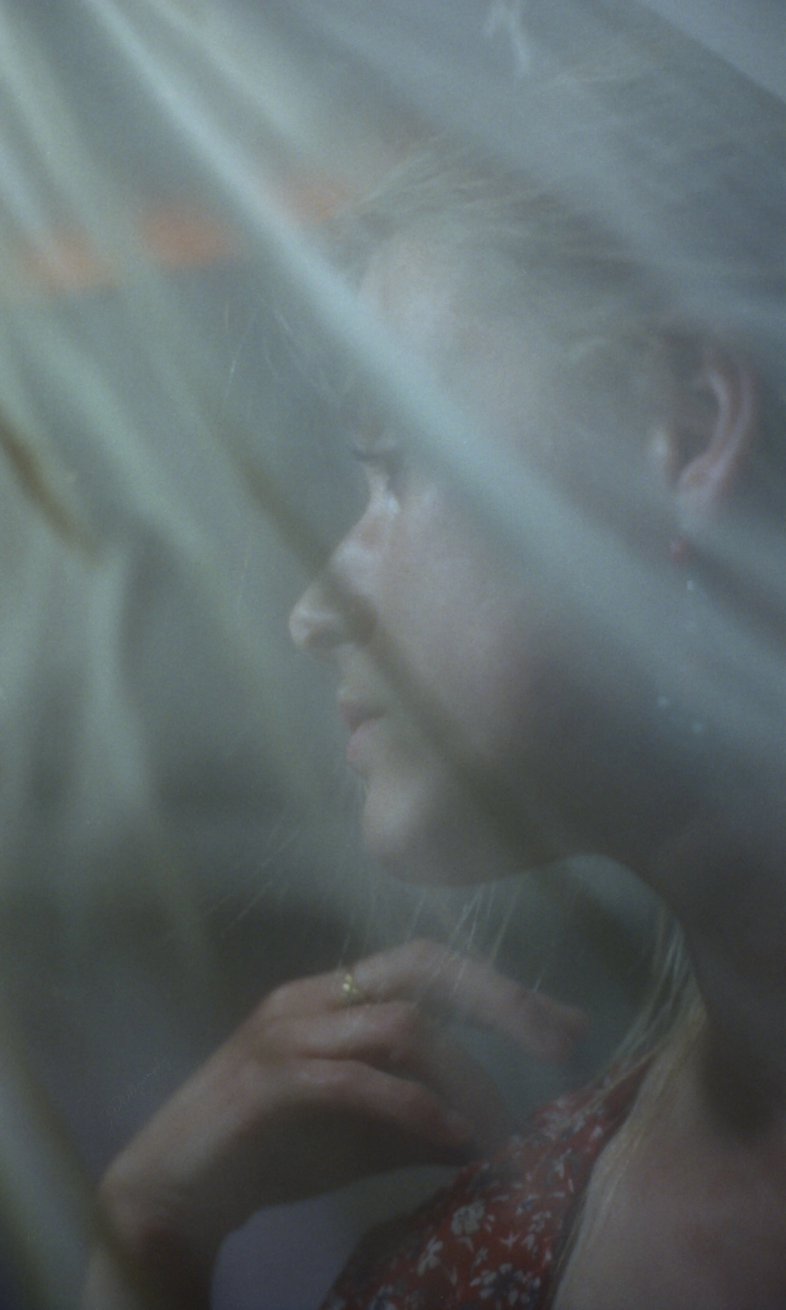 Une jeune femme en profil derrière une surface vitrée ou en plastique, avec ses cheveux blonds et une main portant une bague, portant une robe rouge à motifs blancs.