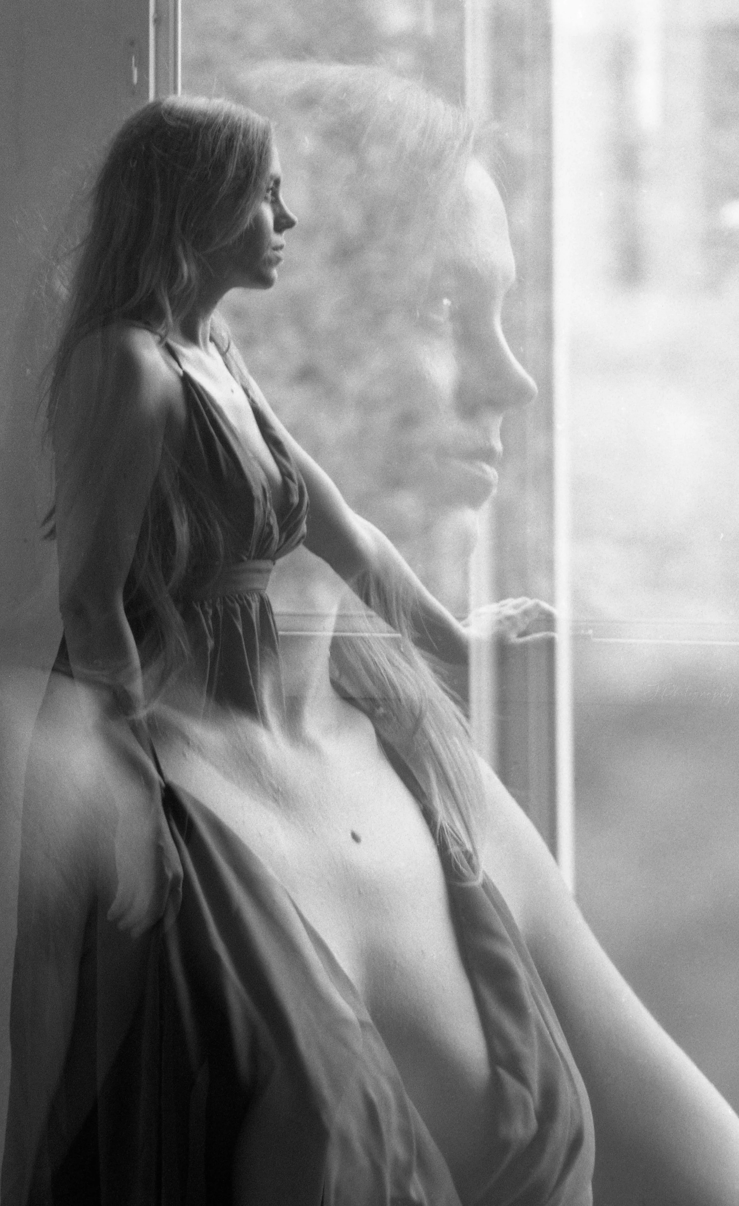 Une double exposition en noir et blanc montrant une femme debout près d'une fenêtre, avec son reflet en gros plan superposé à l'image, créant un effet artistique et contemplatif.
