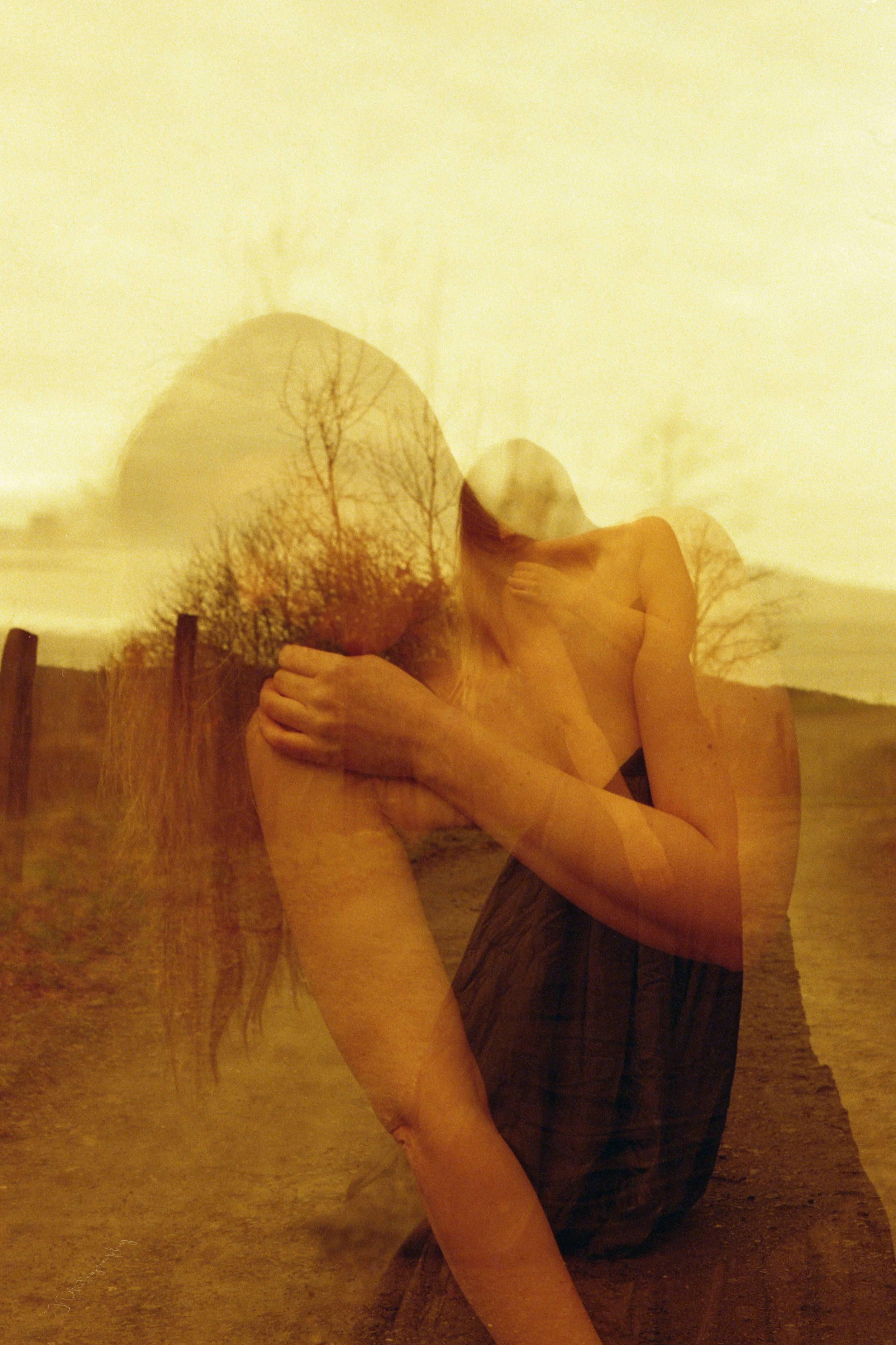 Une femme nue assise à l'extérieur, superposée à un fond de paysage avec des arbres dénudés et un ciel orageux, créant un effet de double exposition