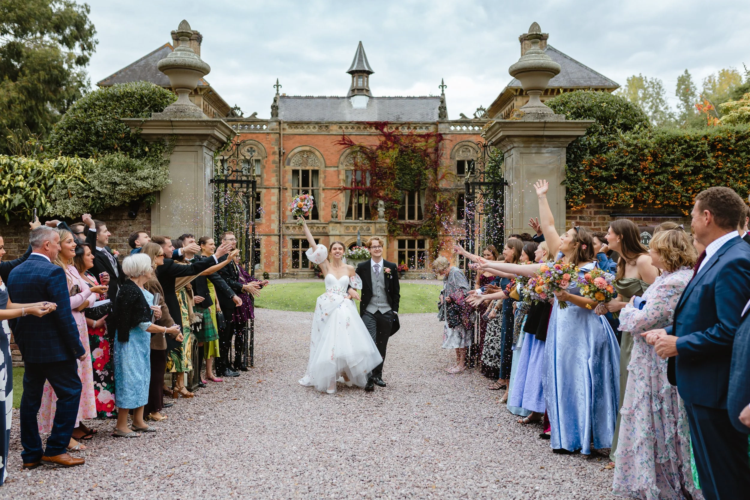 Fun confetti moment outside of Soughton Hall. Bride wears wedding dress with floral details.