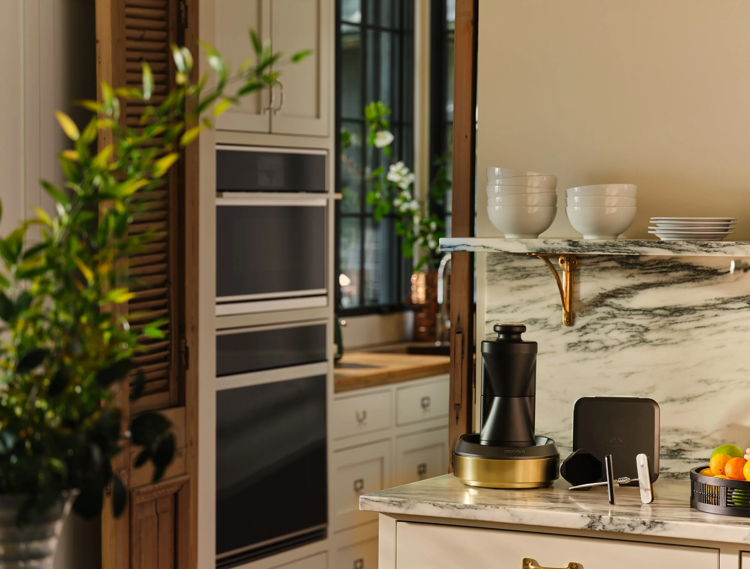 A kitchen counter with a black coffee grinder on a gold base, a small black speaker, and a tray of oranges. Above it, a marble shelf holds white bowls and plates. The background features cabinetry, a window, and greenery.