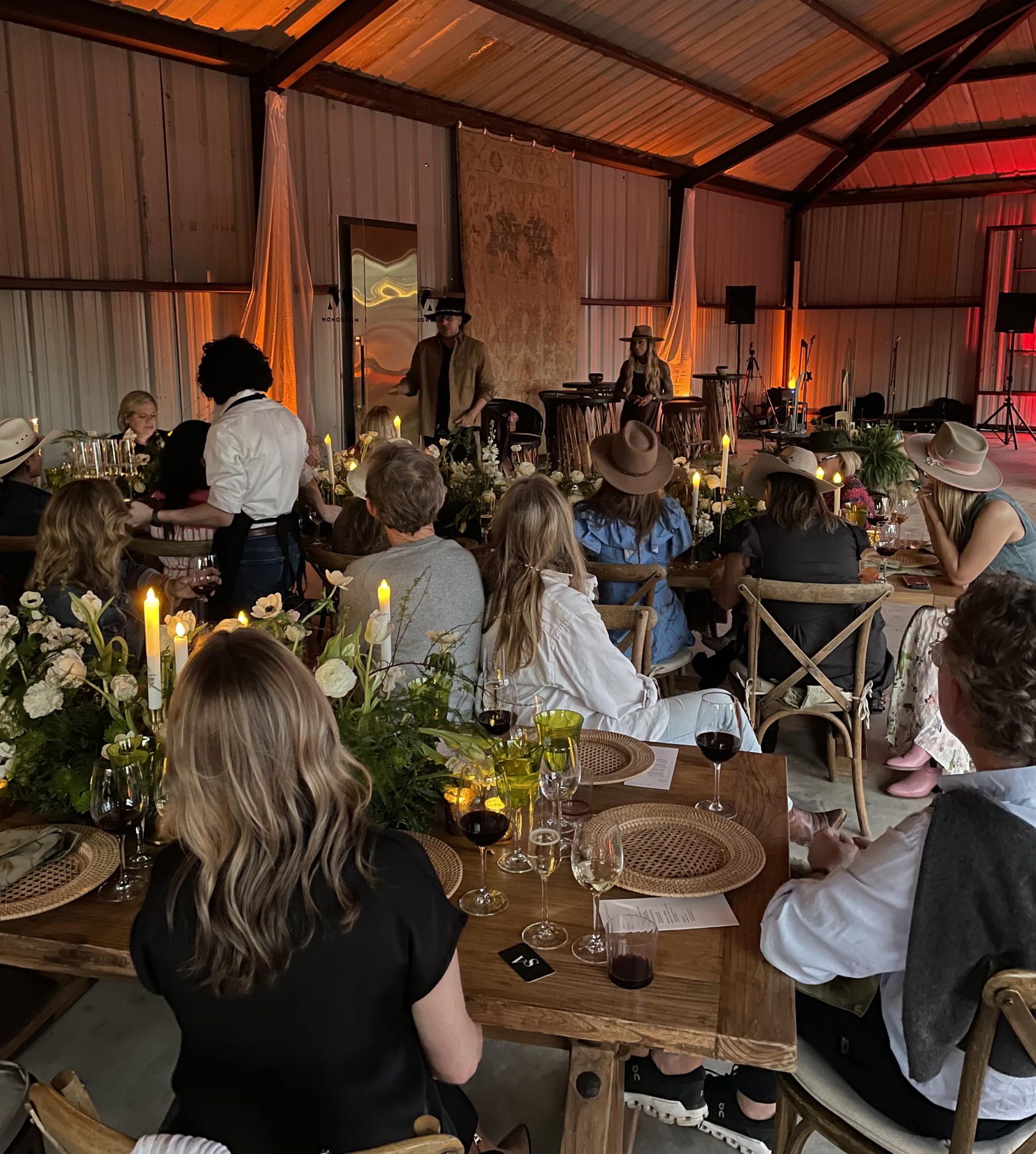 Guests seated at tables with wine glasses and floral centerpieces in a rustic indoor venue watching a live music performance
