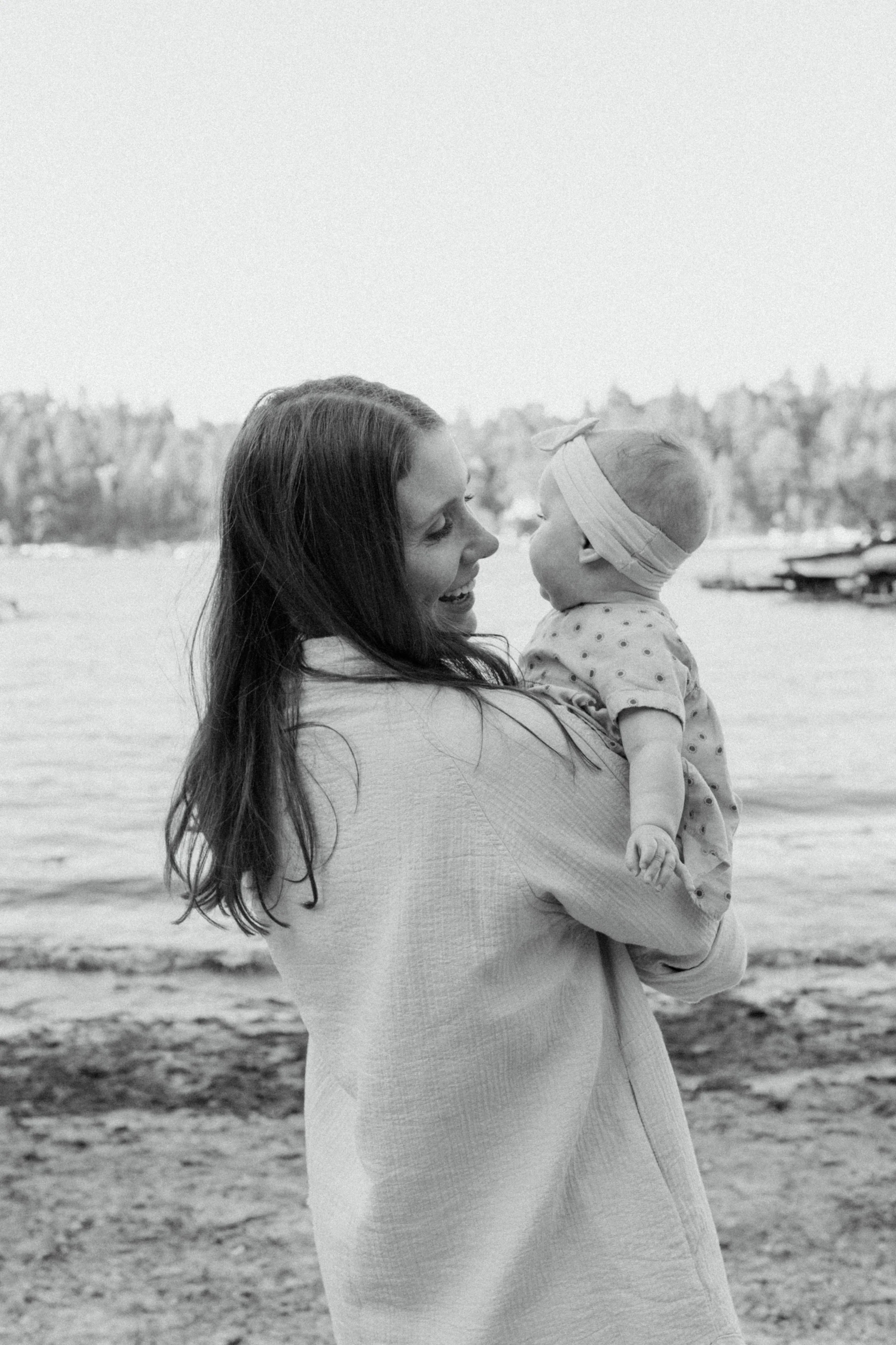 A woman holding a baby while standing on a beach, with water and trees in the background, in black and white.