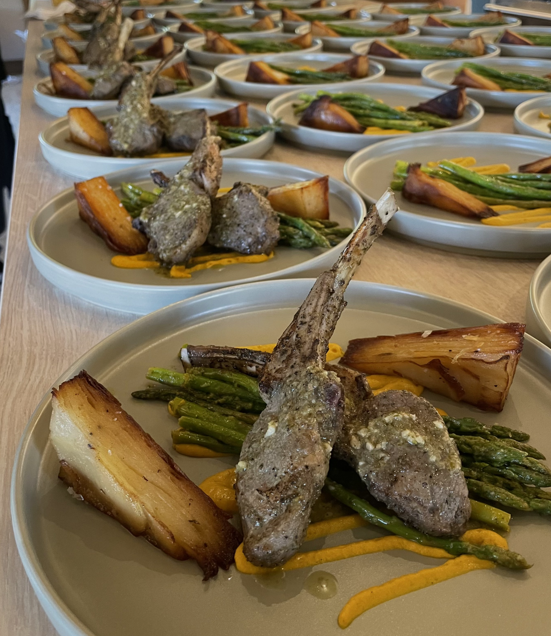 Multiple plates with cooked lamb chops, grilled asparagus, roasted sweet potatoes, and cheesy potato wedges.