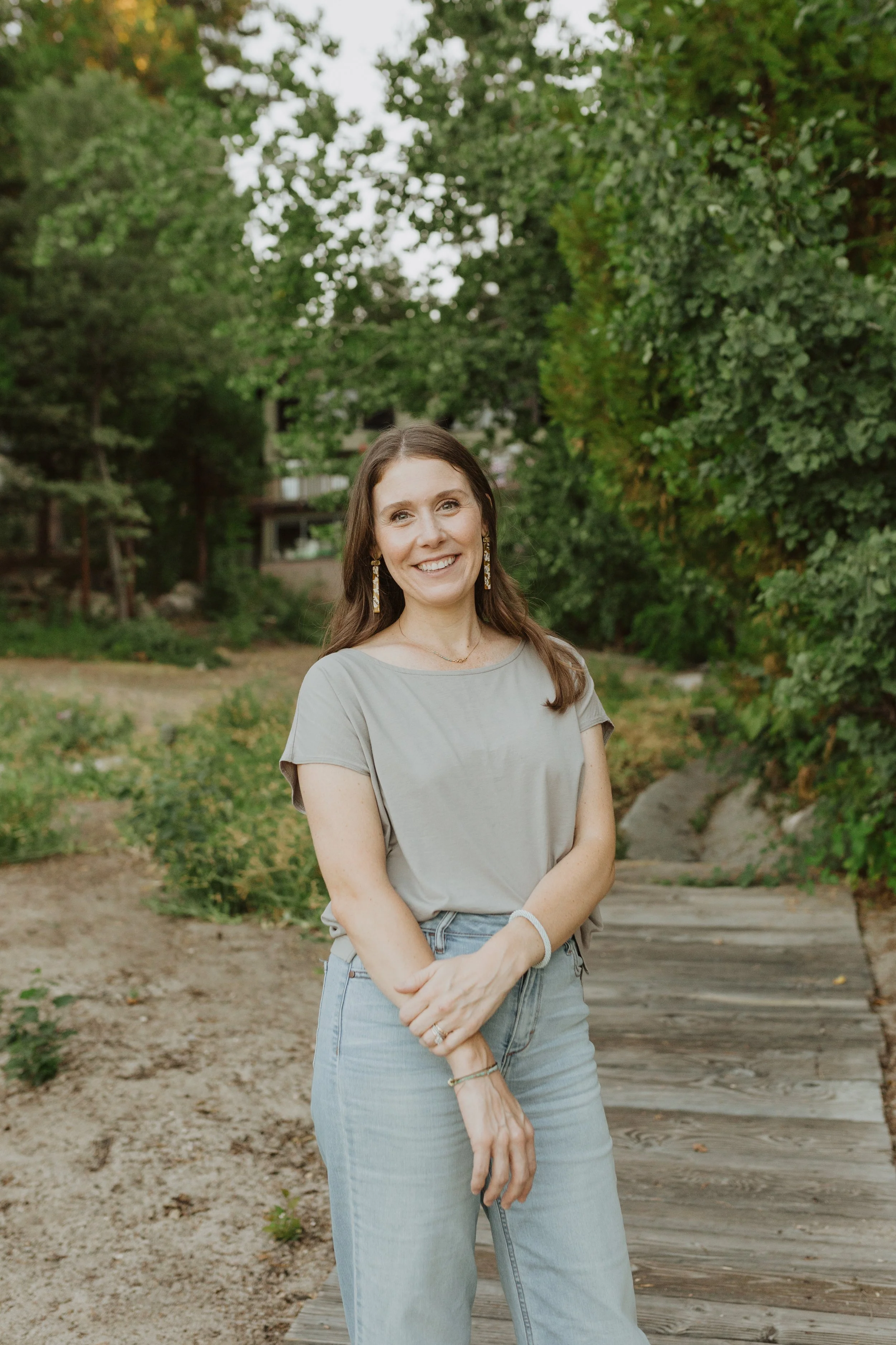 A smiling woman with brown hair, earrings, wearing a light gray t-shirt and jeans, standing outdoors on a wooden pathway surrounded by green trees and bushes.