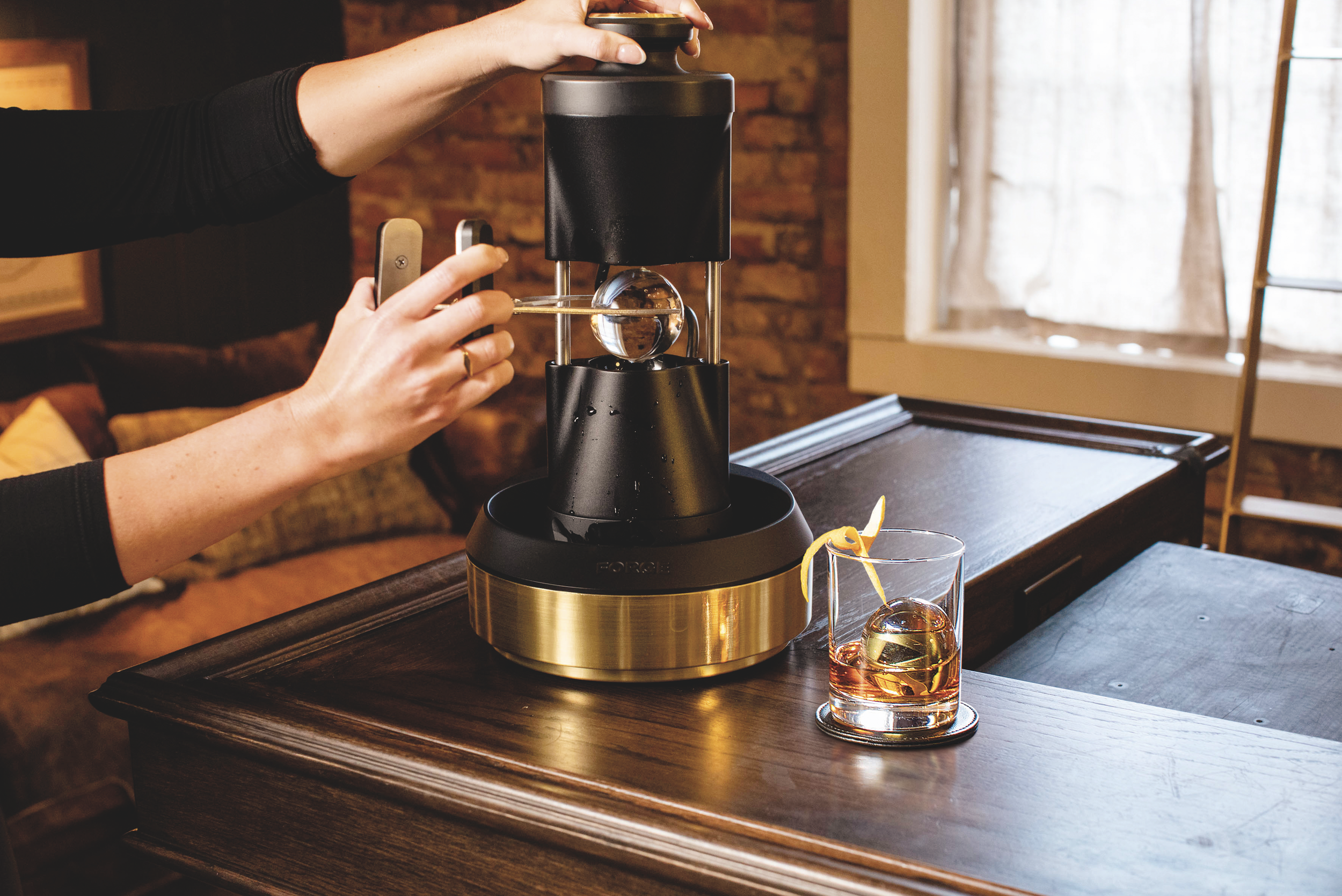 Person using a cocktail sphere ice mold with a defrosting machine on a wooden table, with a glass of an amber-colored beverage garnished with lemon peel.