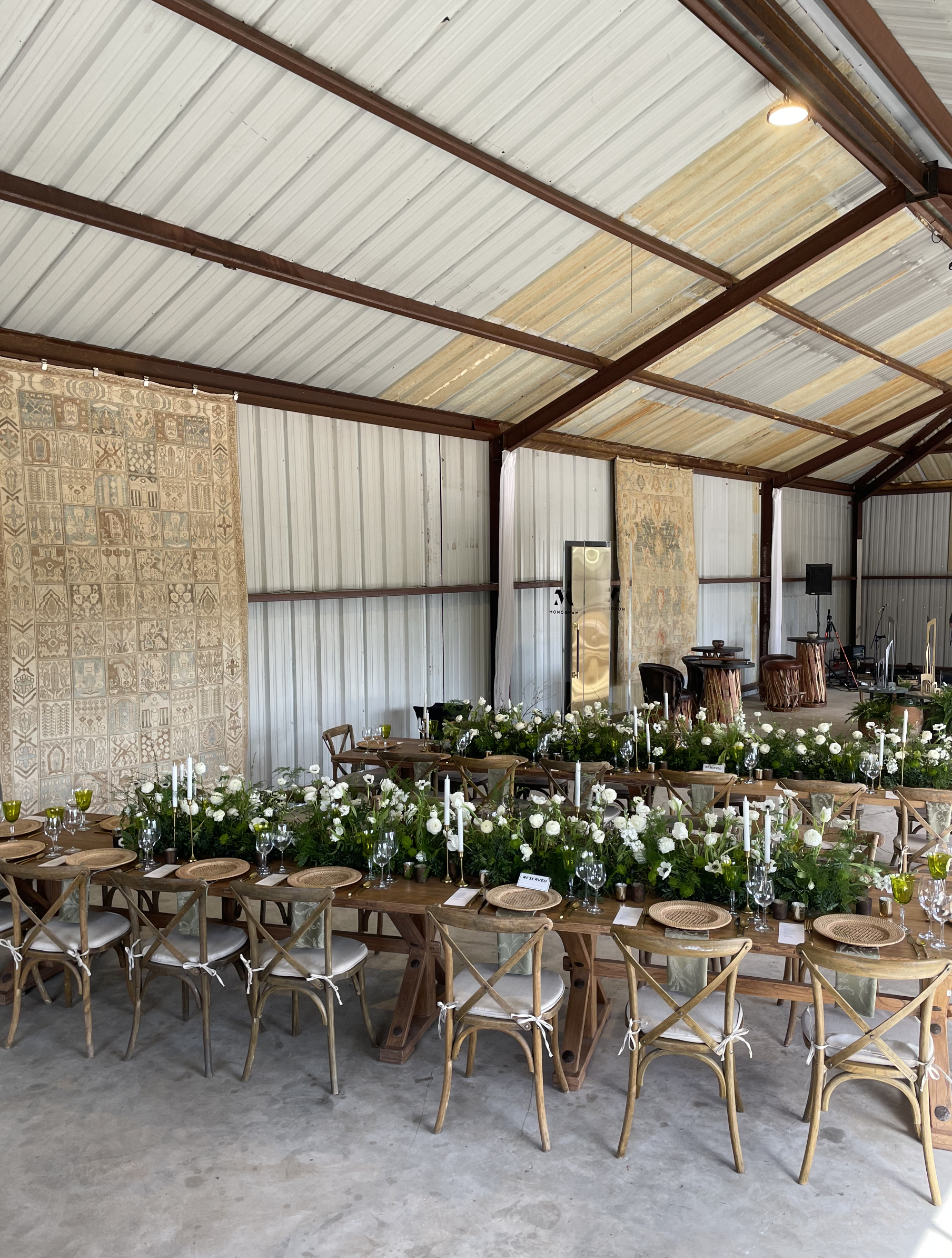 Indoor event space set up for a celebration with a long wooden table decorated with white flowers and tall candles, surrounded by wooden chairs with white cushions, and a backdrop with ornate textiles.