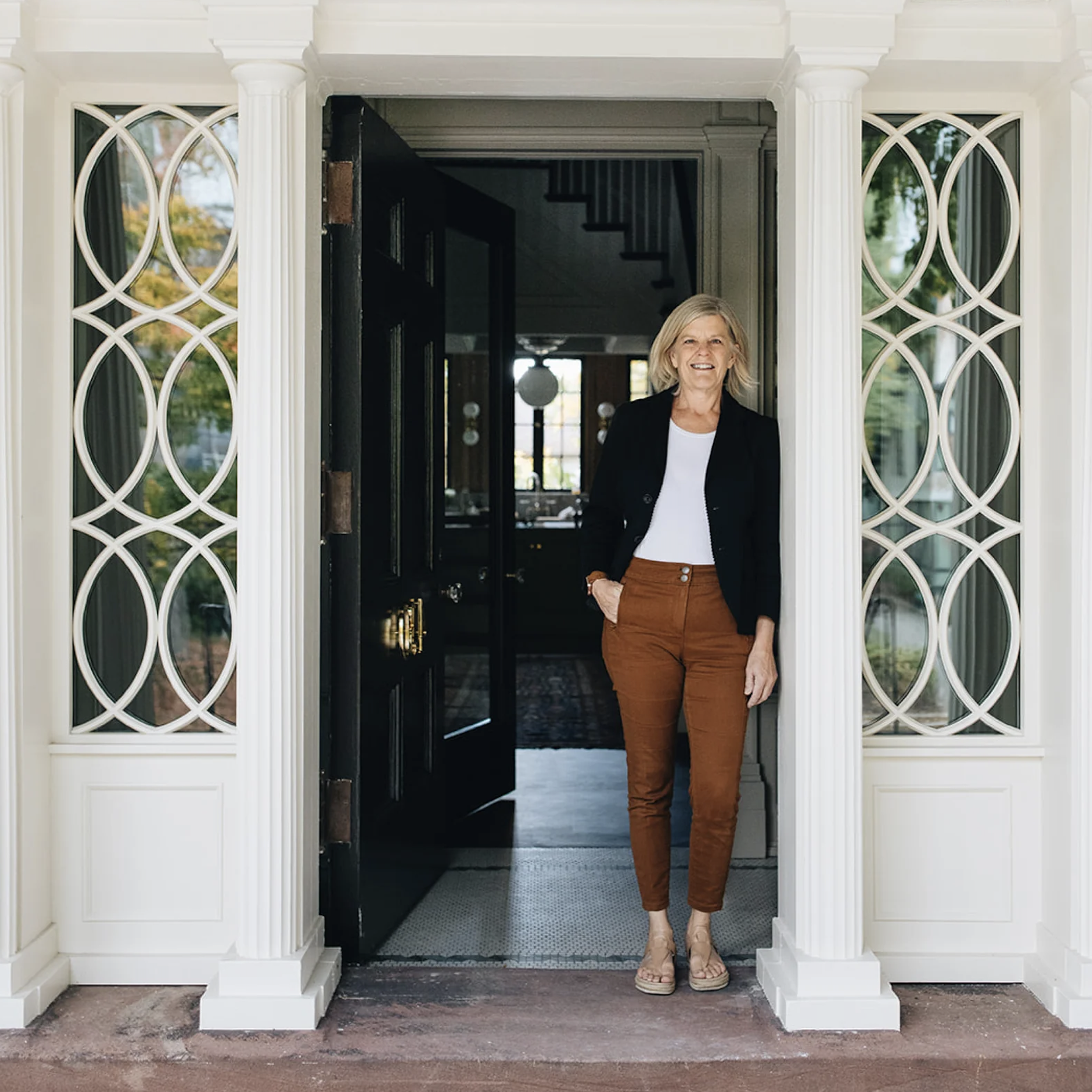 A woman standing at the entrance of her home, smiling, with her hands in her pockets, wearing a black jacket, white shirt, tan pants, and sandals.