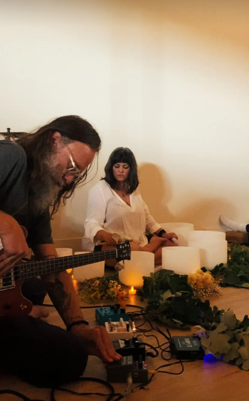 A white woman with dark hair in a white shirt playing crystal singing bowls, sitting next to a white man with long hair and a beard playing bass.