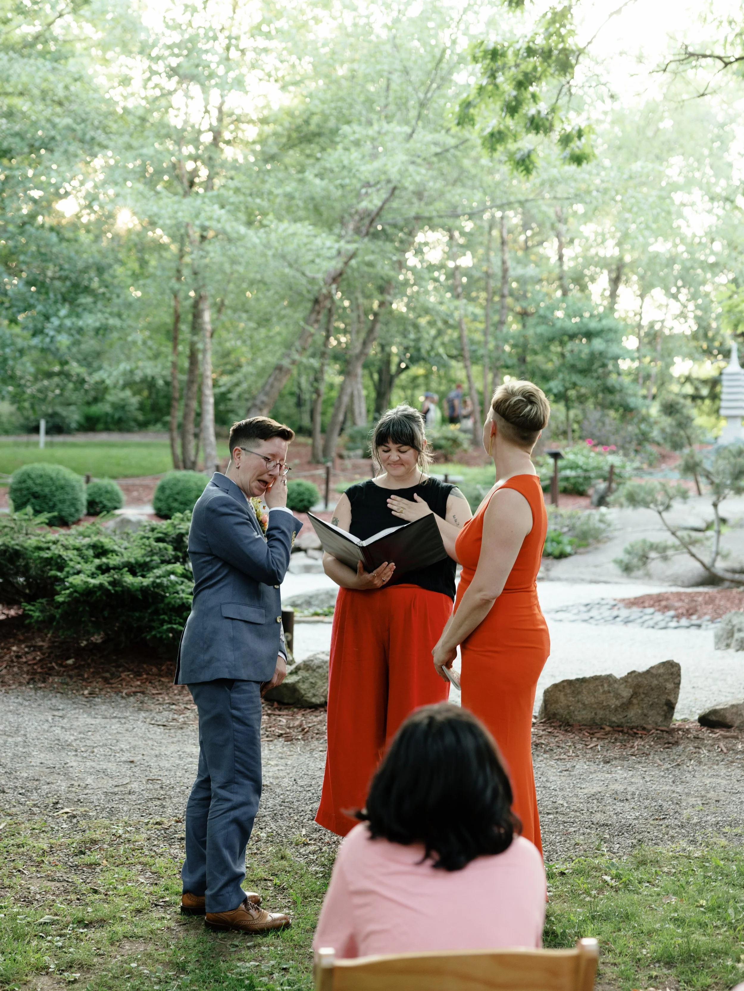 Three people stand under the trees, one in a red dress with blond hair, one in a blue suit with brown hair, and one in red pants and black tank top with gray and brown hair. The one with gray and brown hair is marrying the other two.