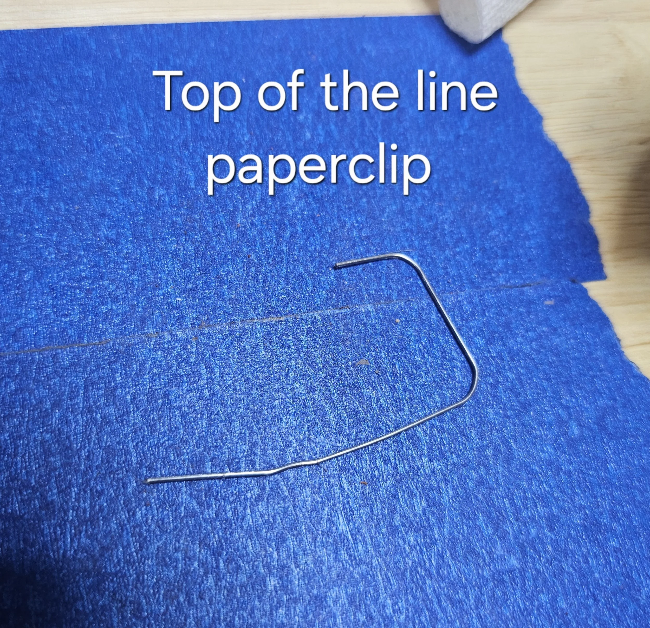 A blue piece of paper with the text 'Top of the line paperclip' printed on it, placed on a wooden surface, with a metal paperclip partially visible.