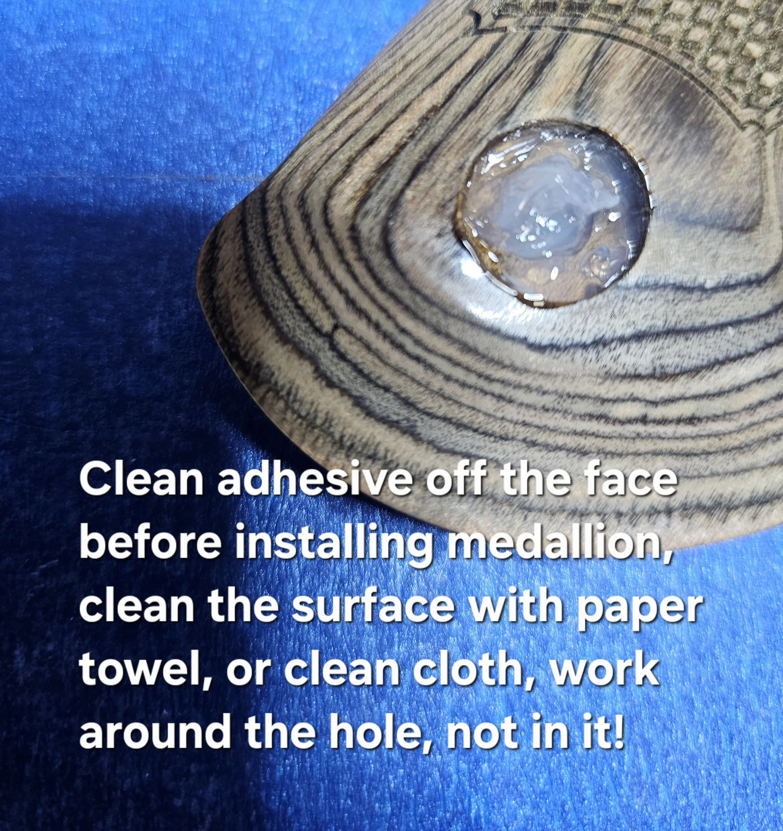 Close-up of a wooden surface with a hole filled with glue, on a blue textured background, with instructional text overlaid that reads: "Clean adhesive off the face before installing marquee, towel, or clean cloth, work around the hole, not in it!"
