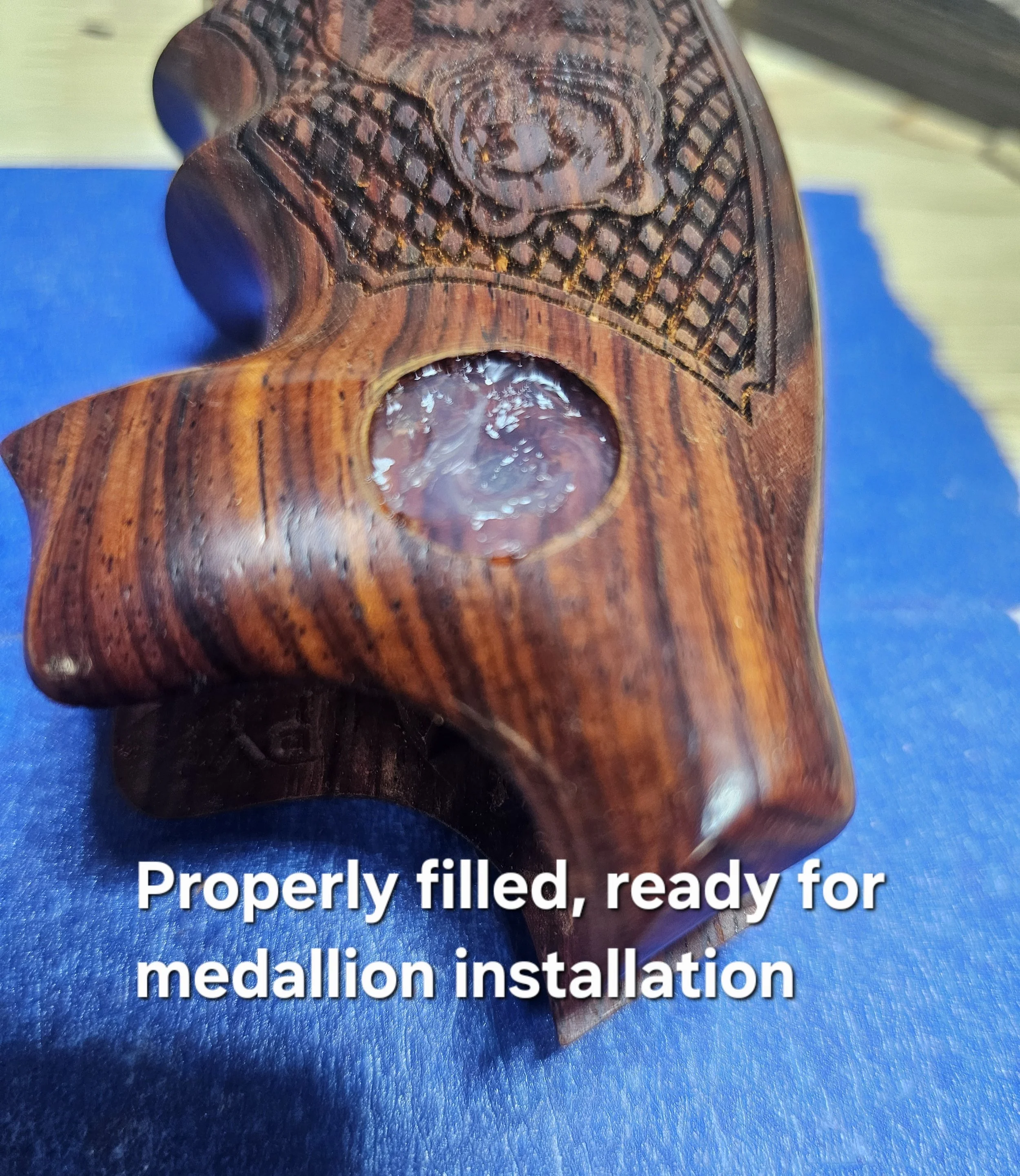 Close-up of a wooden medal plaque with engraved text, placed on a blue surface, with a partial view of a metallic medal at the bottom.