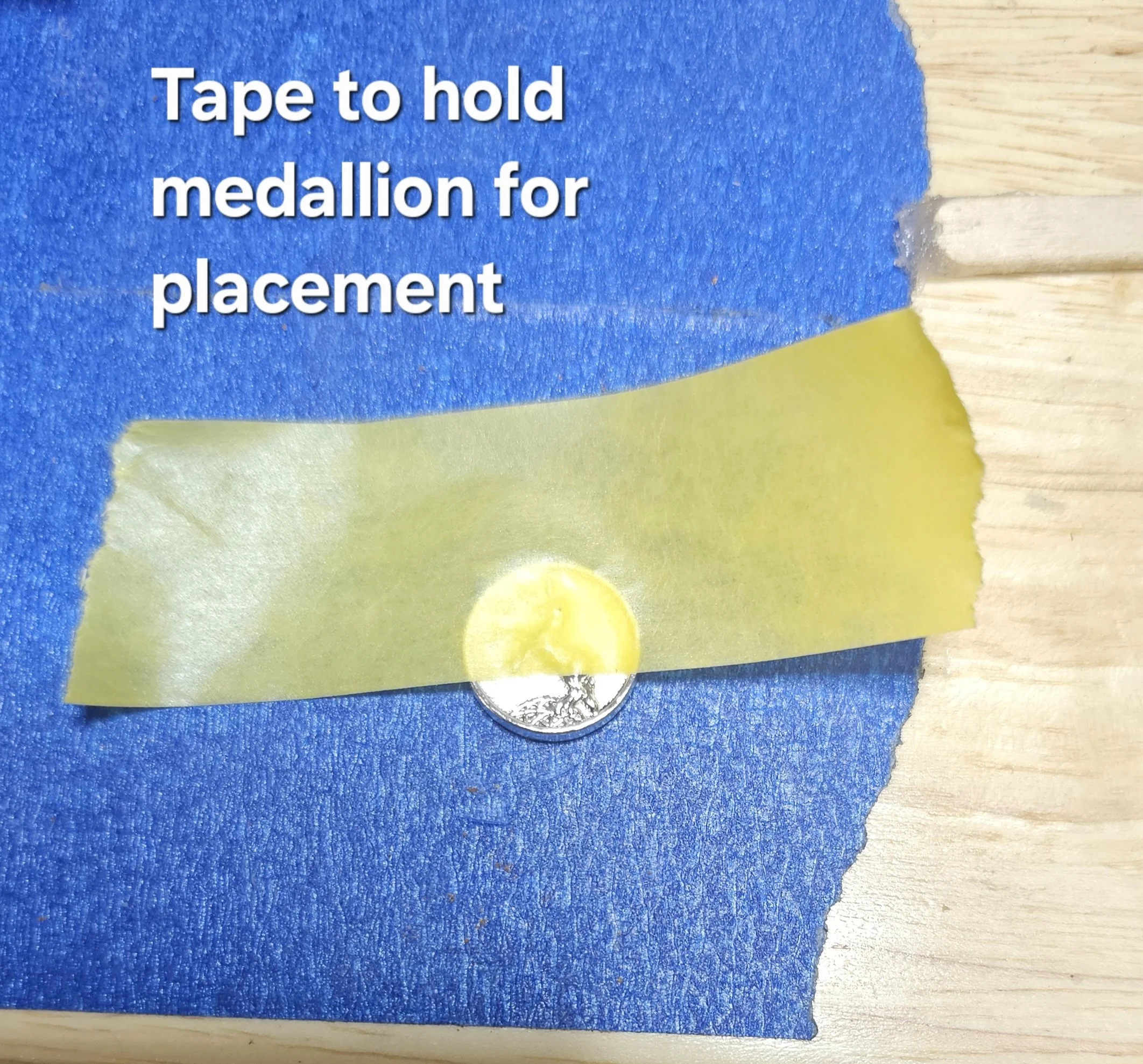 Close-up image showing a piece of blue painter's tape, a yellow sticker, and a silver coin on a light-colored wooden surface with some torn blue tape.