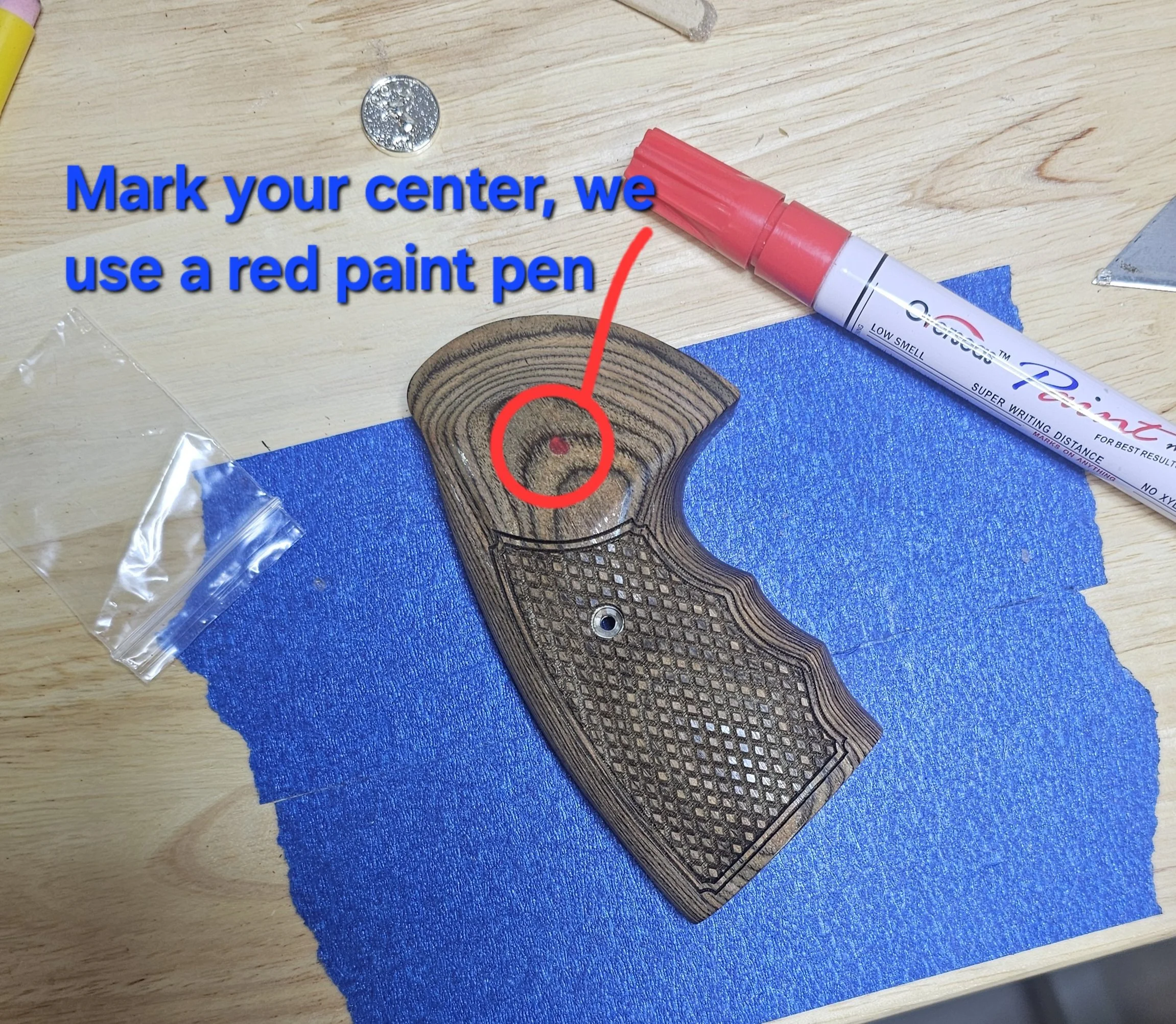A wooden textured object with a small circular hole, placed on a blue textured cloth. There is a pink marker and a metallic piece nearby on a wooden surface. Text over the image instructs to mark the center of the object using a red paint pen.