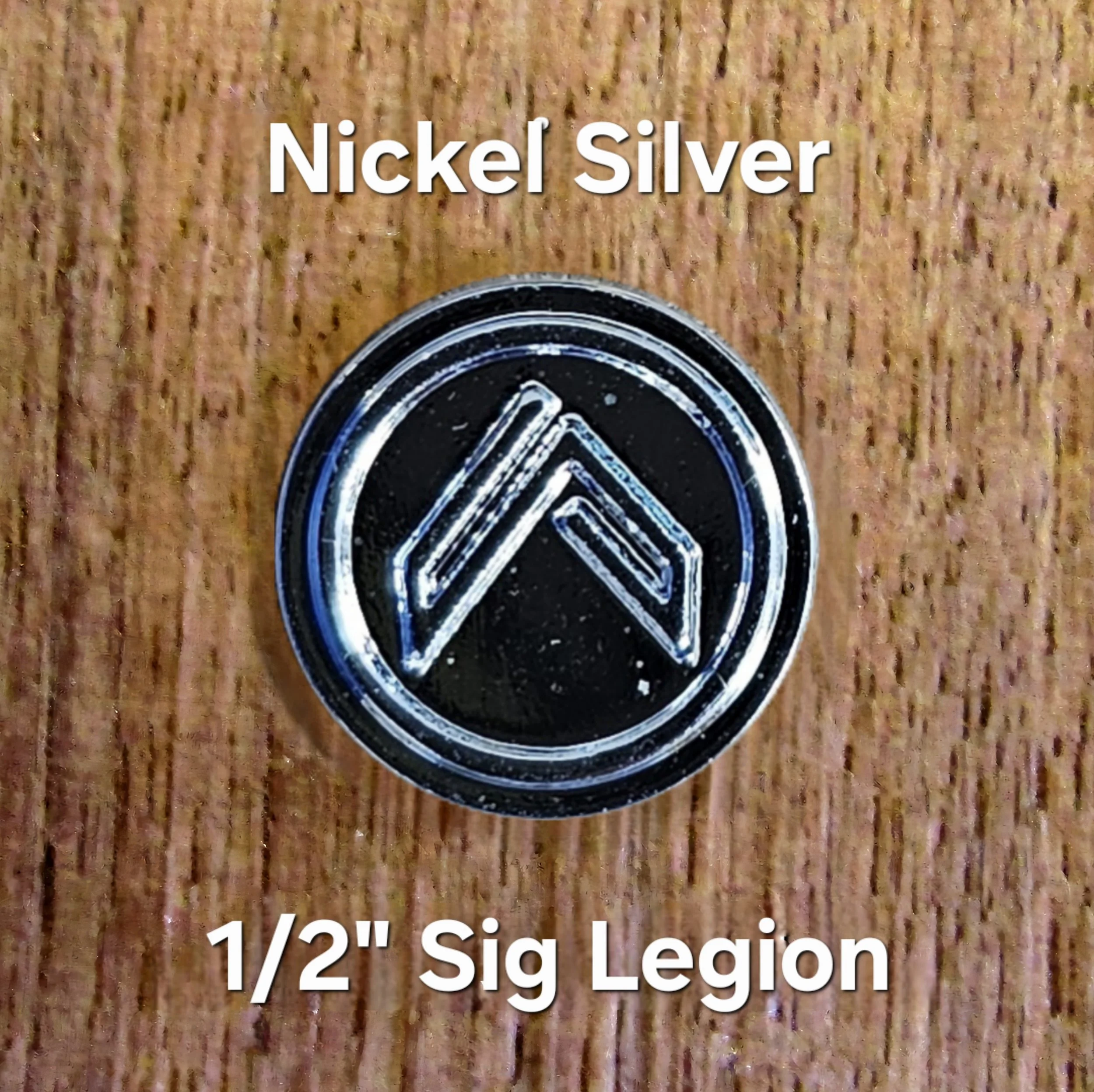 Close-up of a black and white coin with a circular design, placed on a wooden surface. Overlaid text states 'Nickel Silver' and '1/2" Sig Legio'.