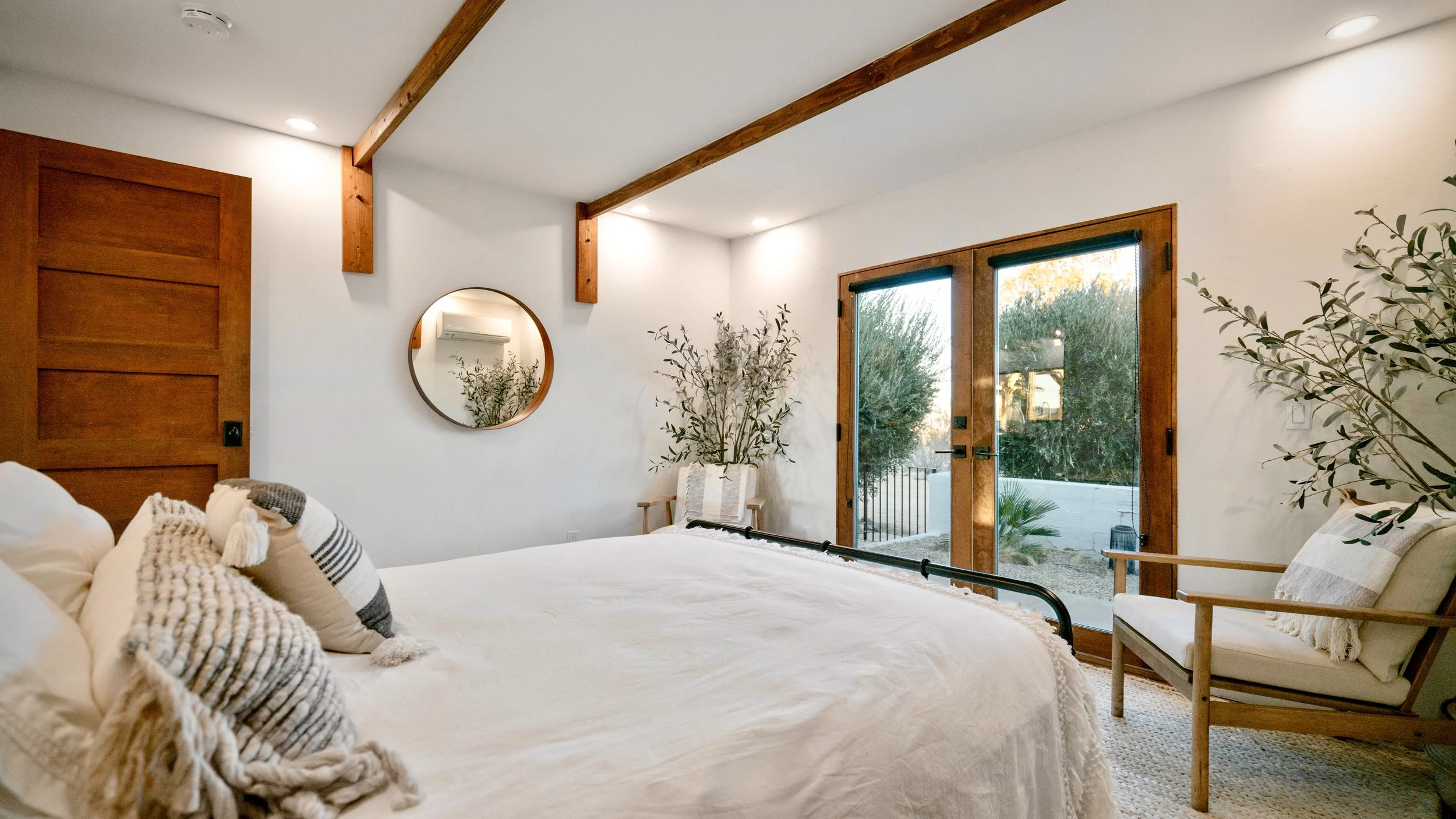 View across the full-size bed in Bedroom 3 toward French doors, lounge chairs, a round mirror, and desert landscaping outside.