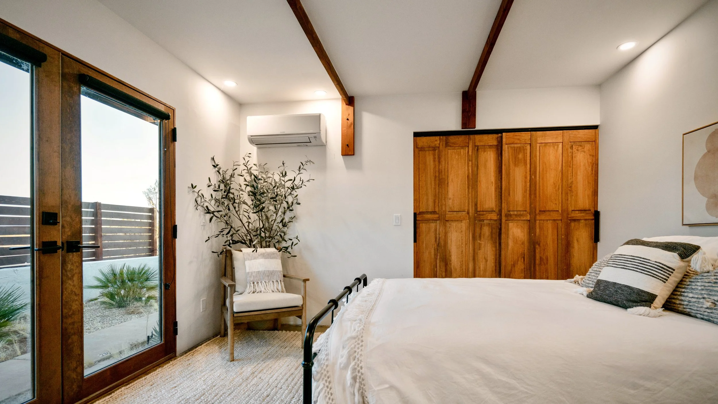 Alternate view of Bedroom 3 showing the full-size bed, built-in wooden closet, reading chair, mini-split, and French doors.