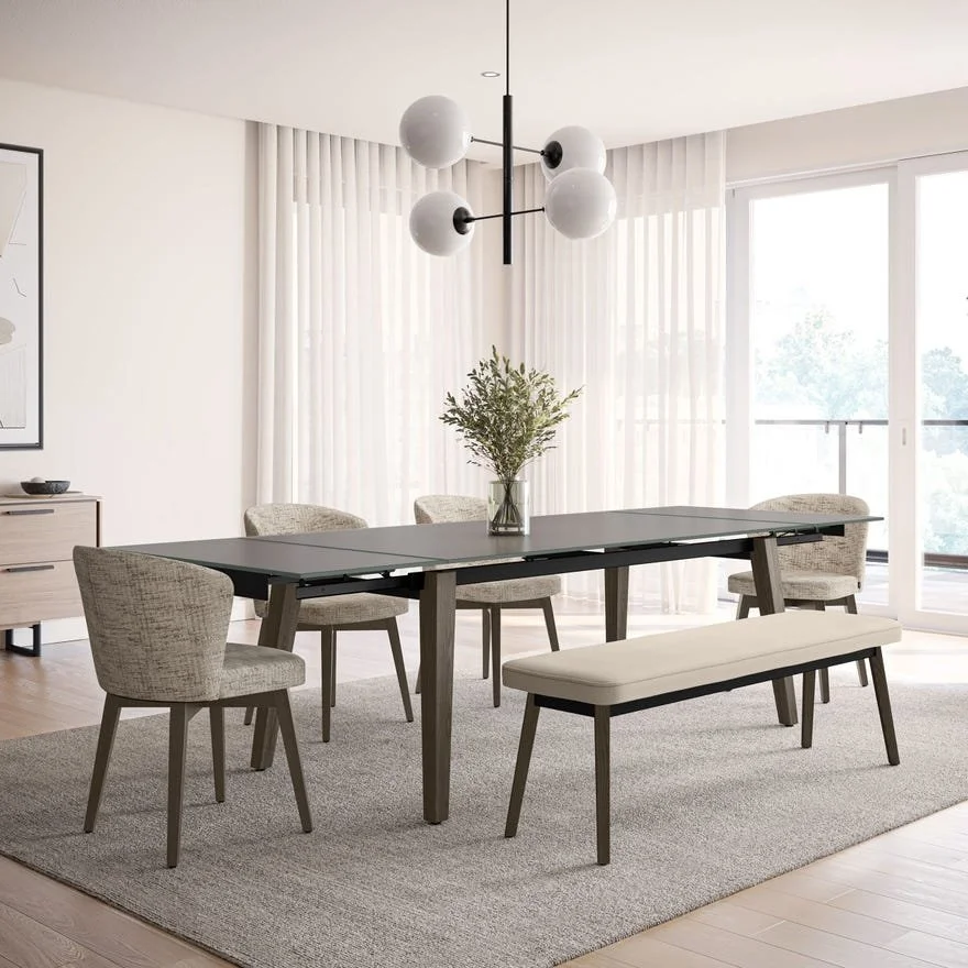 Modern dining room with a black table, six beige upholstered chairs, a beige bench, a gray rug, a glass vase with greenery on the table, sheer white curtains, and large sliding glass doors leading to a balcony.