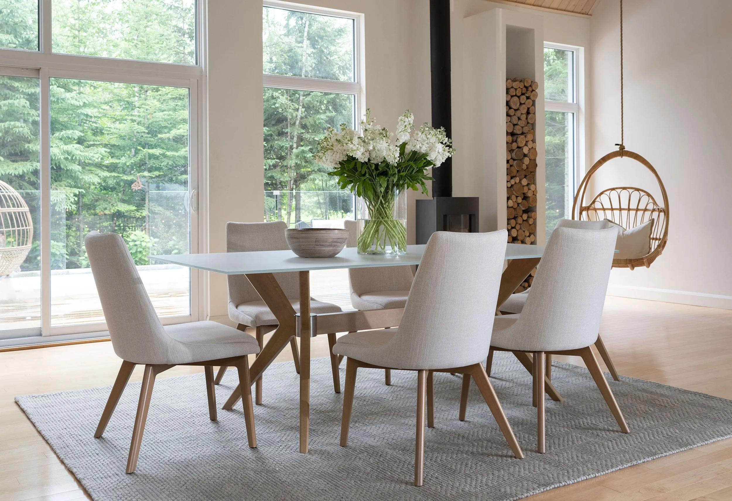 Modern dining room with a white table, six cream-colored chairs, a vase with white flowers, a bowl on the table, large windows showing greenery outside, a wood-burning stove, and a hanging papasan chair.
