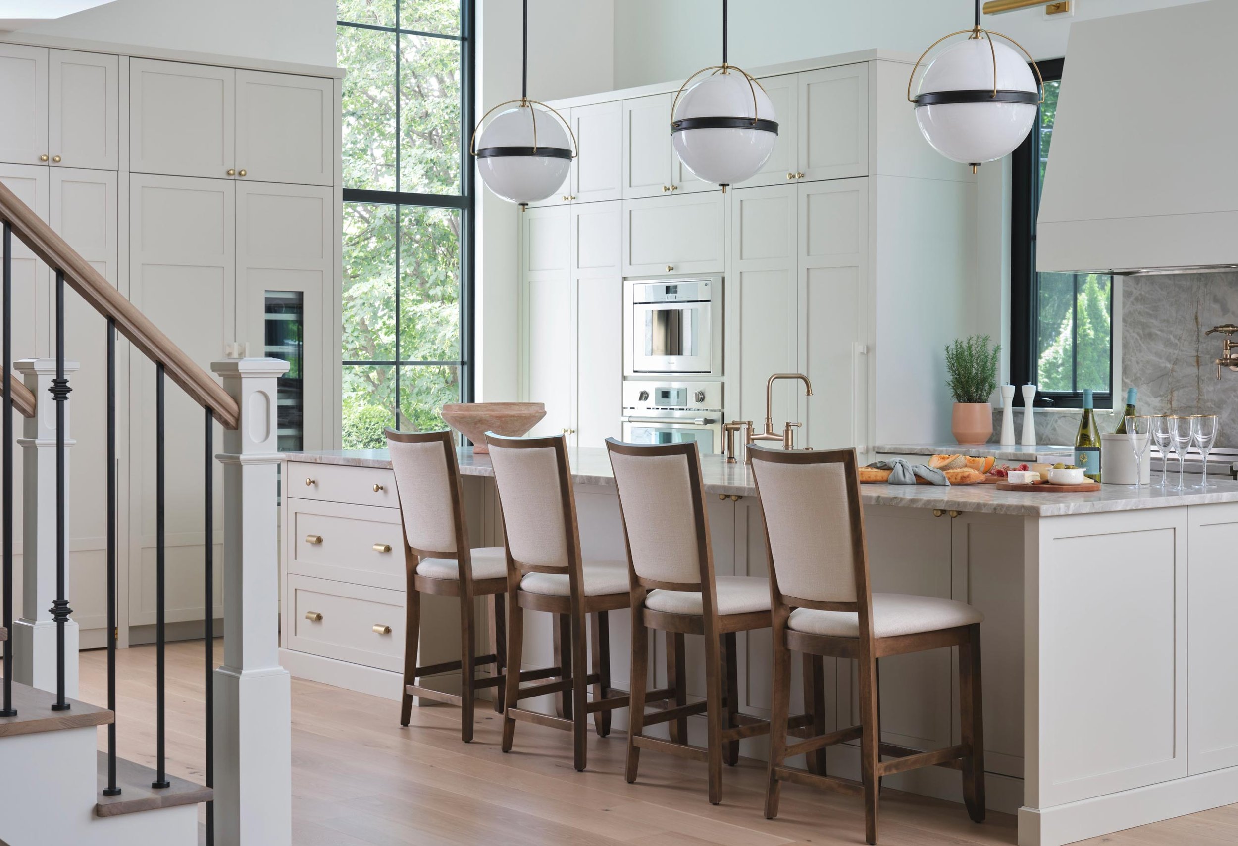 Modern kitchen with white cabinetry, marble countertops, and large windows, featuring a breakfast bar with five wooden chairs and three hanging globe pendant lights.