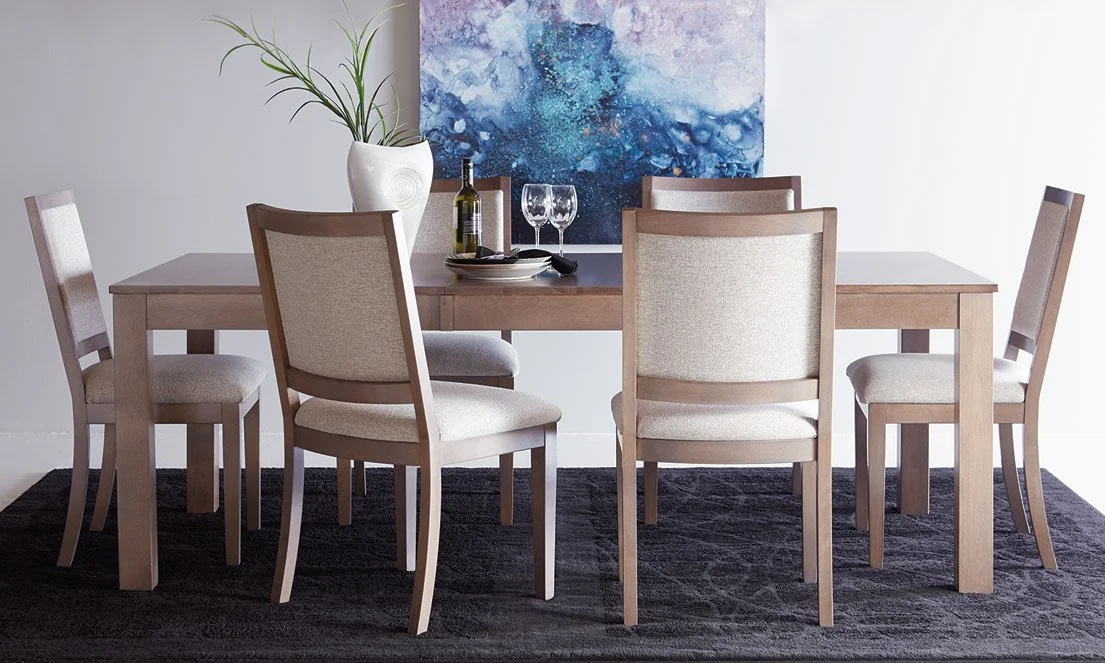 A dining room table with six beige upholstered chairs, set with a wine bottle, two wine glasses, and some plates, with a potted plant and abstract blue artwork in the background.