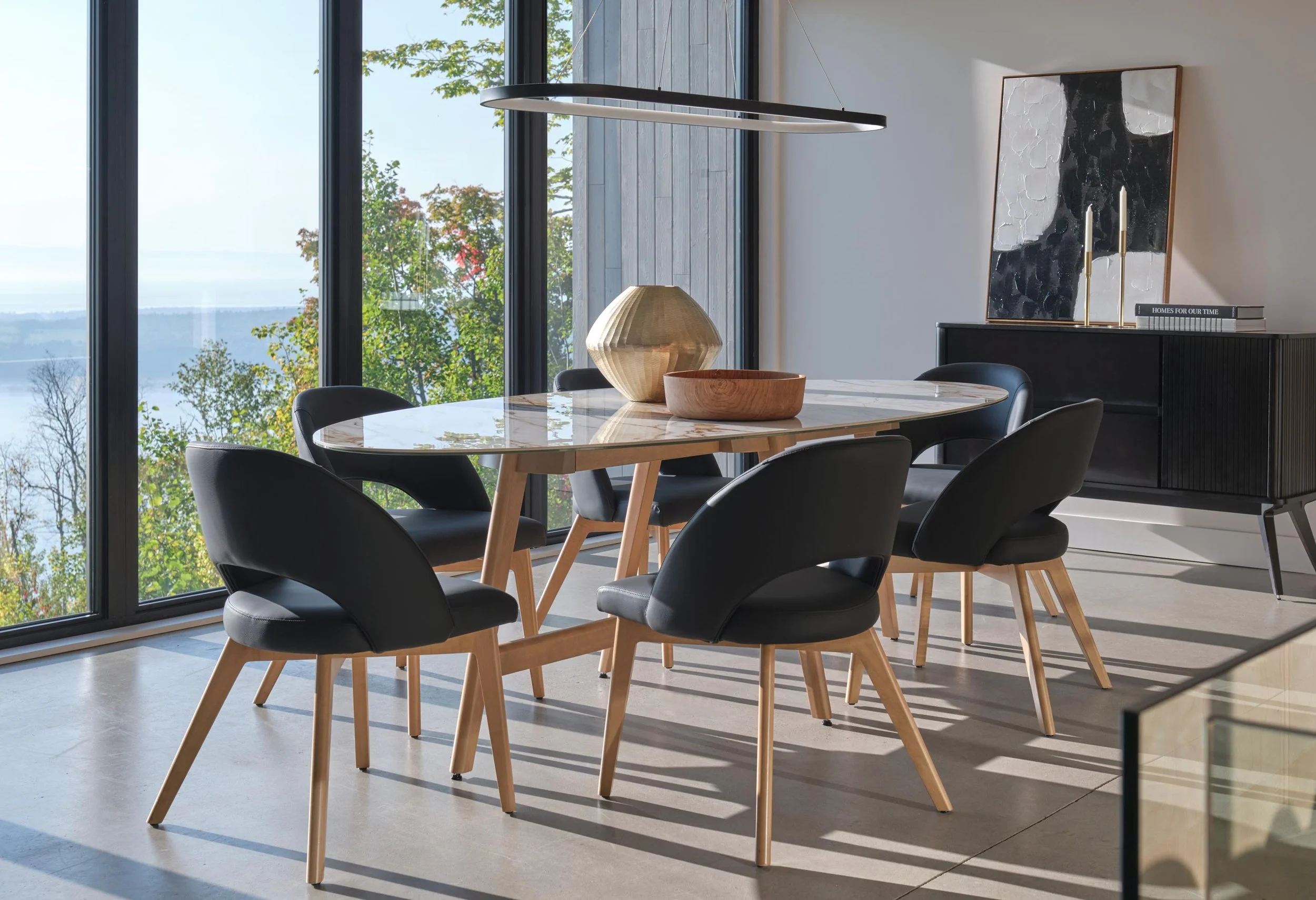 Modern dining room with six black chairs with wooden legs around a marble-top table, large glass windows with a view of trees and water outside, and a black sideboard with decorative items and a framed abstract painting.
