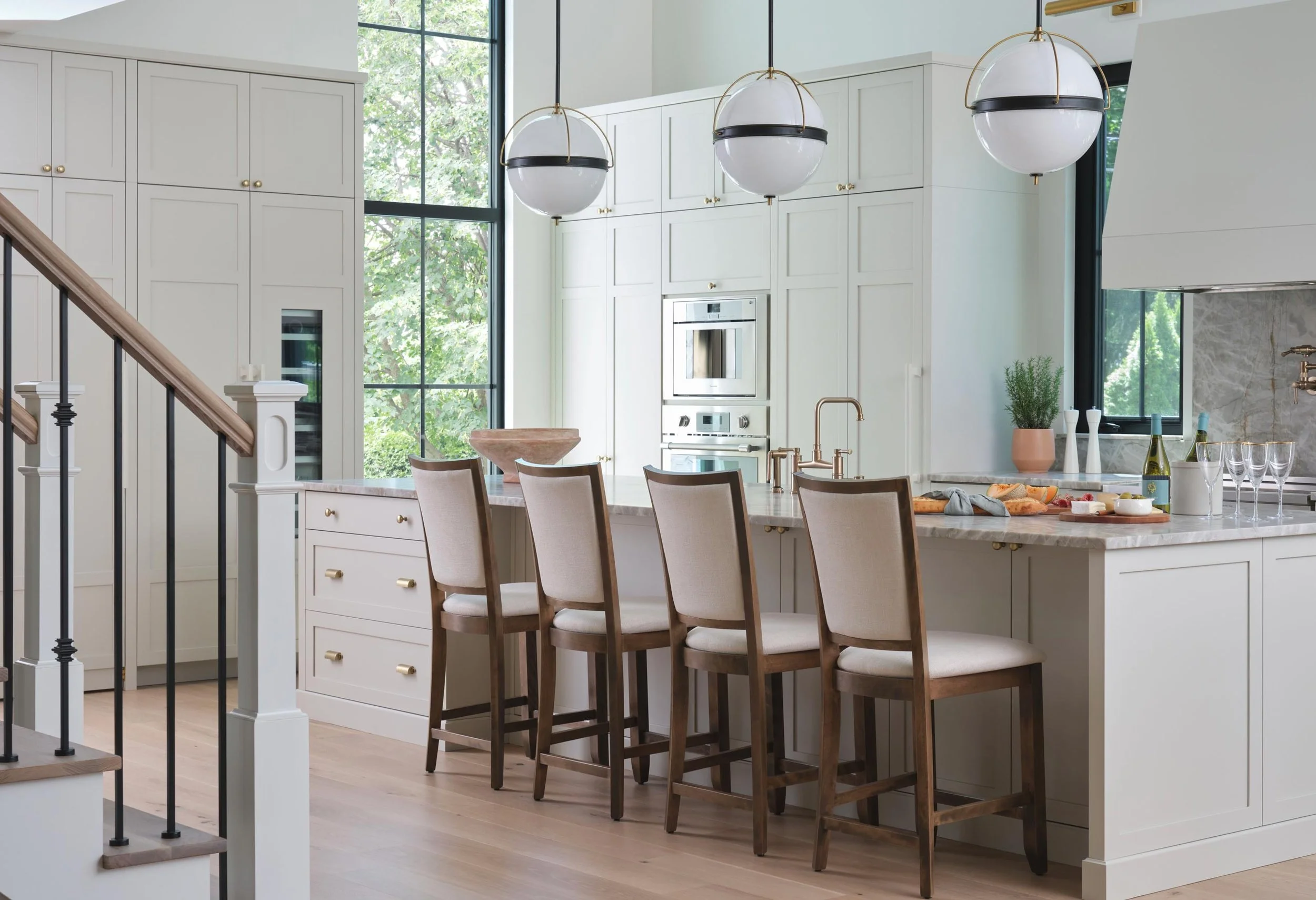 Modern kitchen with white cabinetry, large window, marble island, four beige chairs, hanging pendant lights, and various kitchen items.