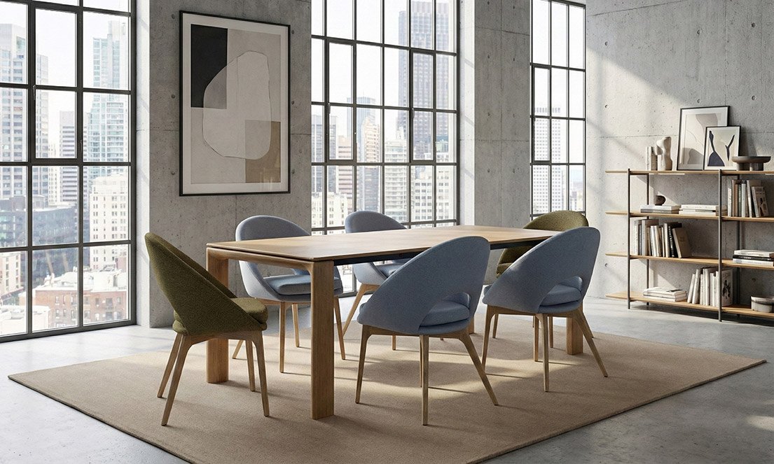 Modern dining room with a wooden table, six upholstered chairs in blue and green, a large abstract art piece on the wall, concrete walls, large windows with city view, and a bookshelf with books and decorative items.