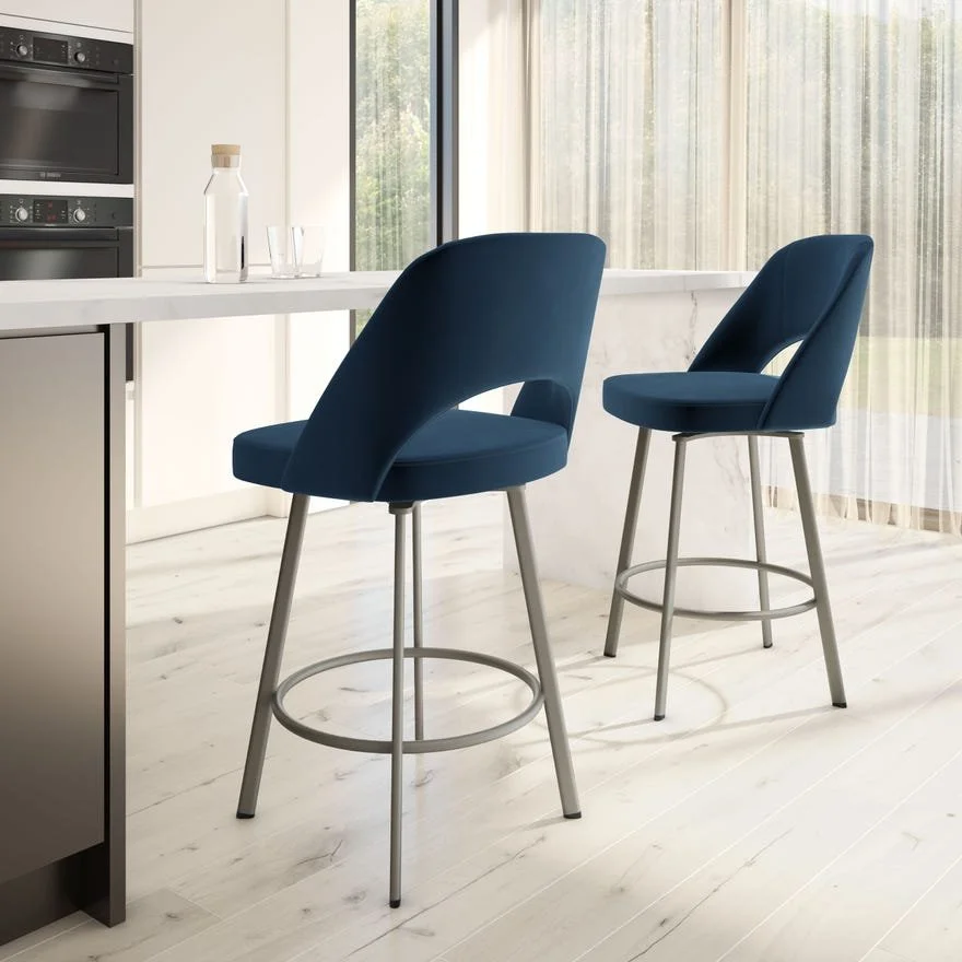 Modern kitchen with two blue bar stools at white marble counter, sunlight through large windows.