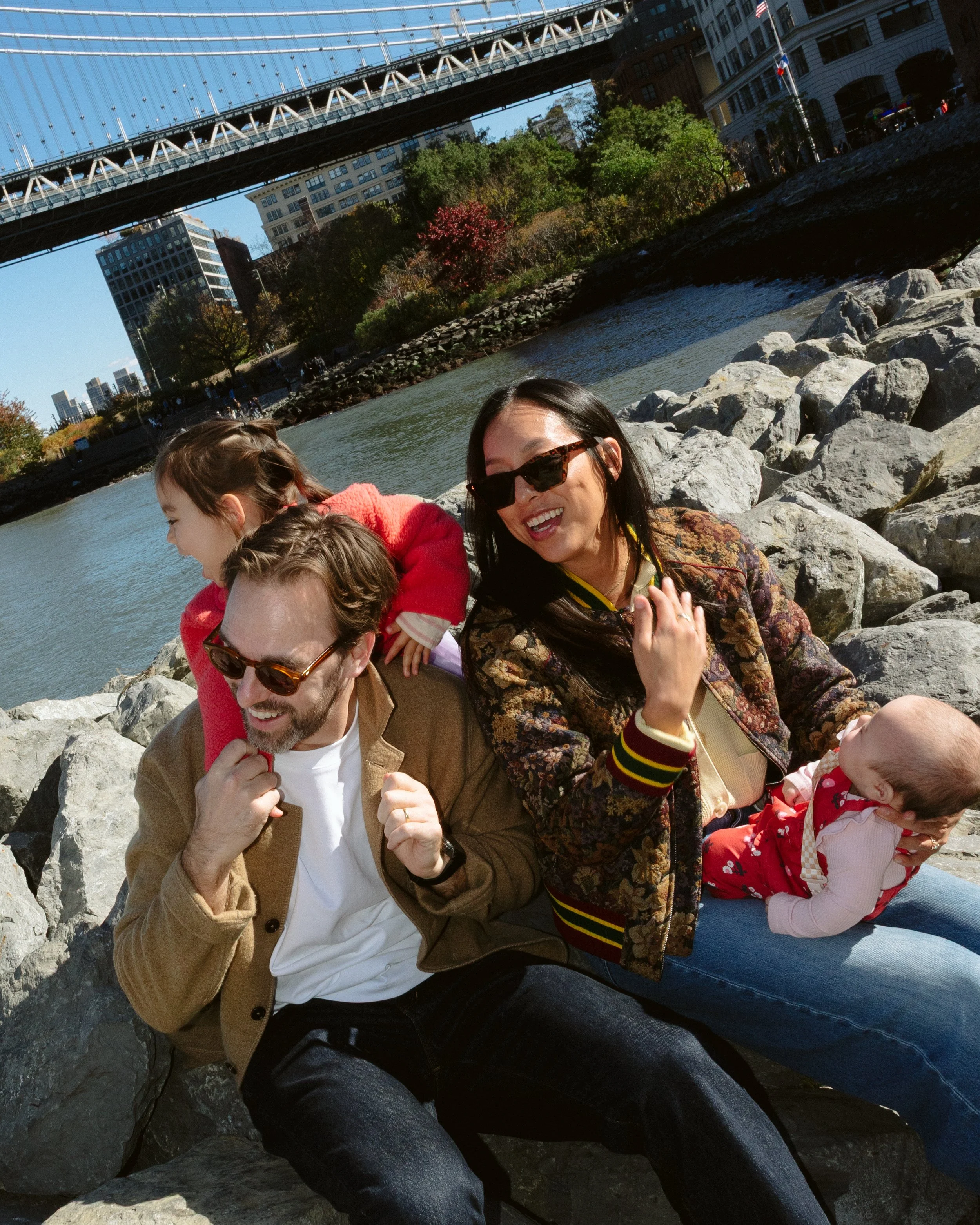 A family of four, including two adults and two children, sitting on rocks near a river with a bridge overhead in New York City, enjoying a sunny day.