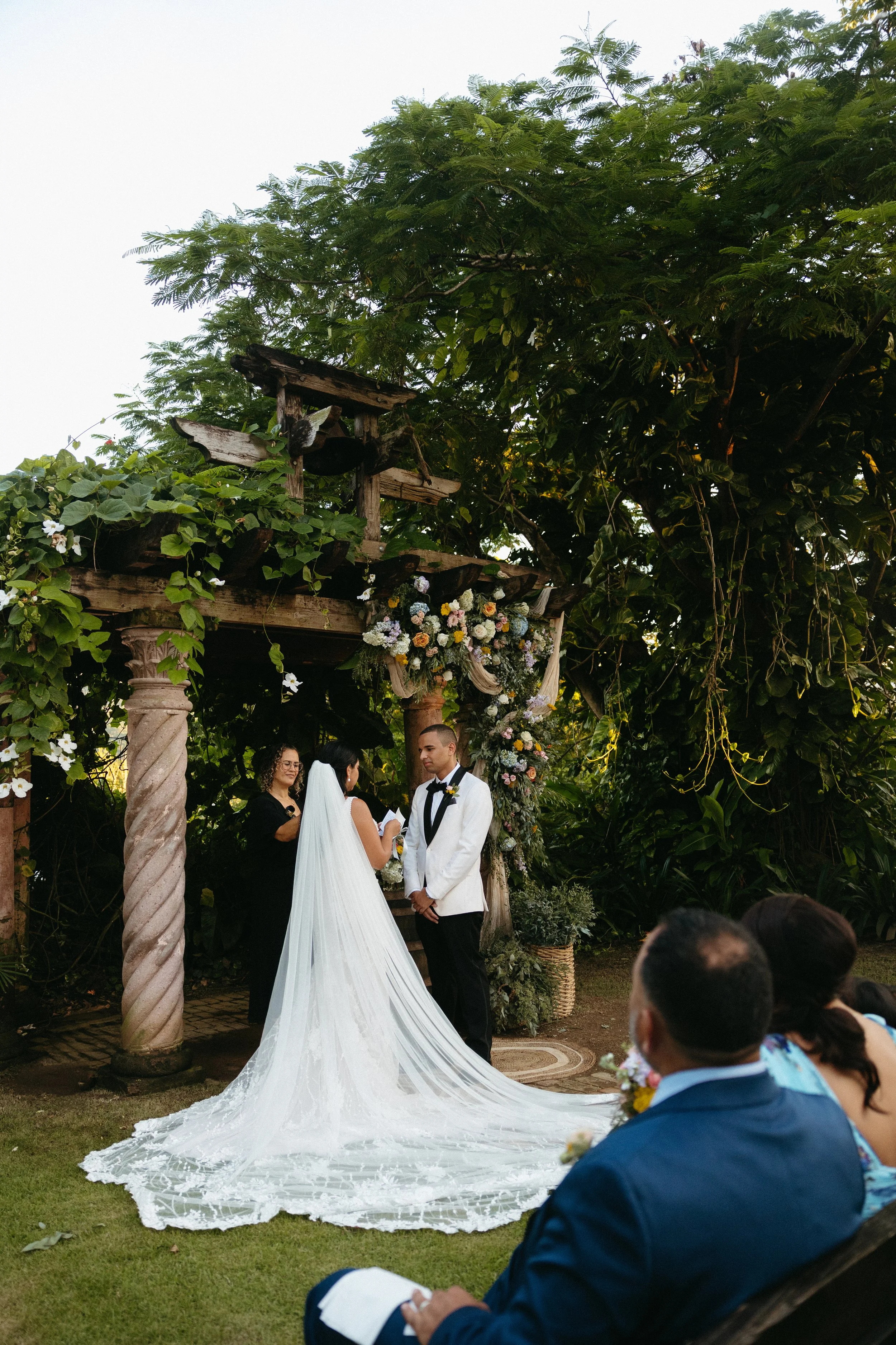 A wedding ceremony taking place outdoors in puerto rico under a wooden, vine-covered arch with floral decorations. The bride and groom stand facing each other, with an officiant standing beside them. Guests are seated on benches.