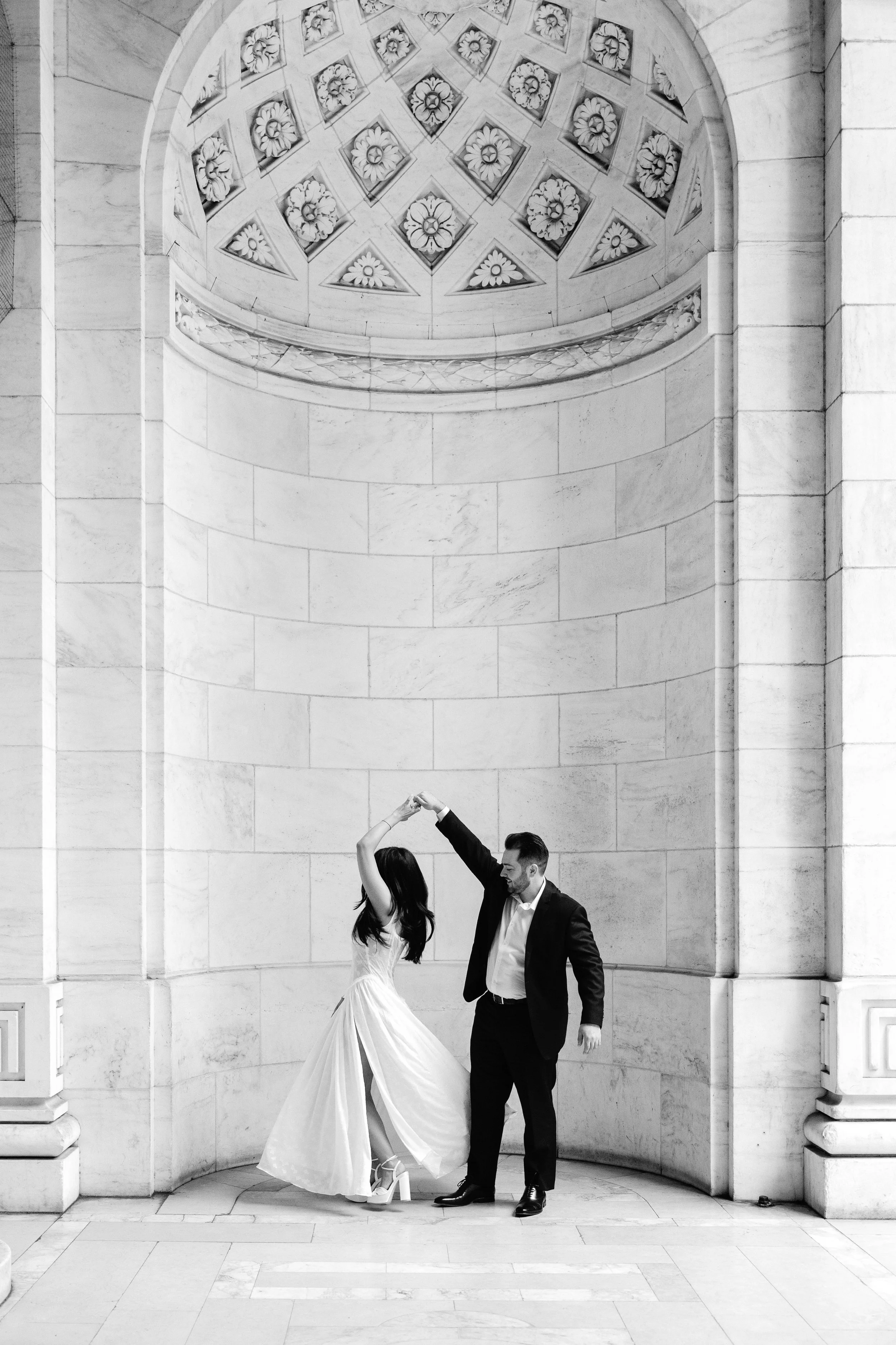 A couple dancing in a grand, marble-beamed hall with an ornate, circular stained glass window above them at the New York Public Library in NYC.