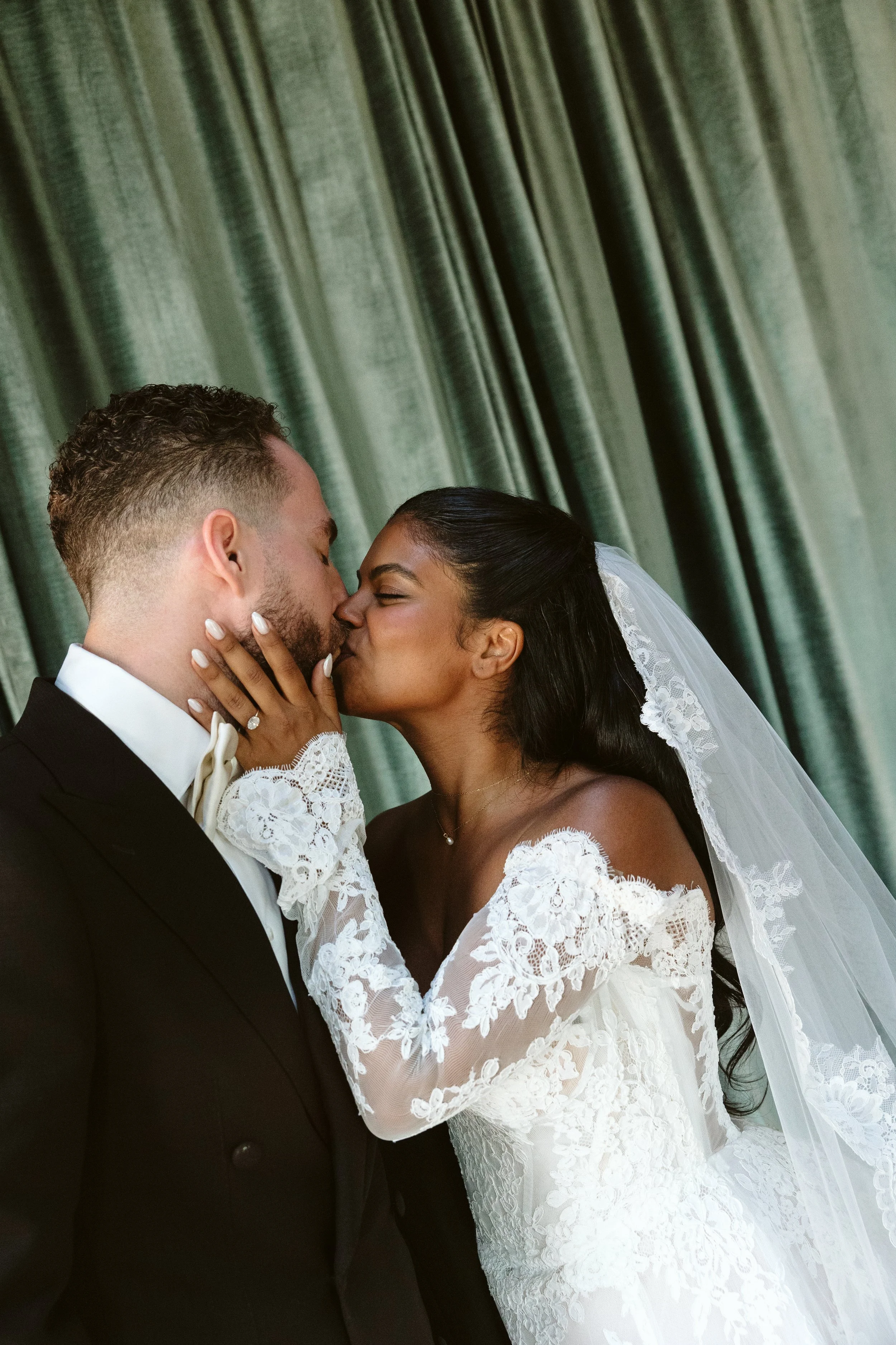 A bride and groom kiss in front of green curtains during their wedding.